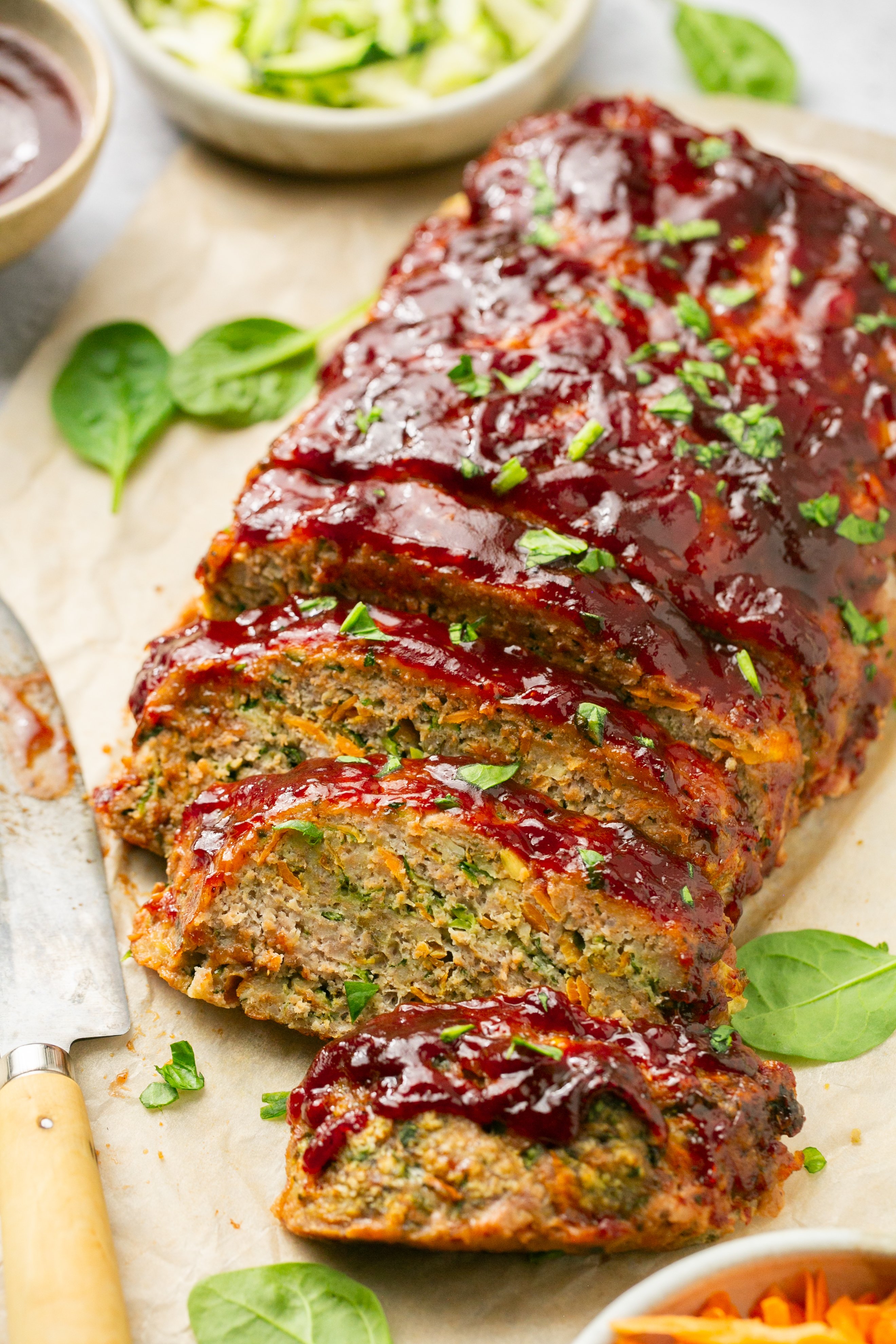Sliced meatloaf covered with barbecue sauce and flecks of spinach on top. Meatloaf is sitting on  parchment paper on a counter with a few small spinach leaves surrounding the meatloaf; strewn about on the parchment paper. Knife with barbecue sauce is sitting on the counter next to the meatloaf; out of frame. Three small bowls are set to the side out of frame. The small bowls contain grated carrot, grated zucchini, and barbecue sauce.