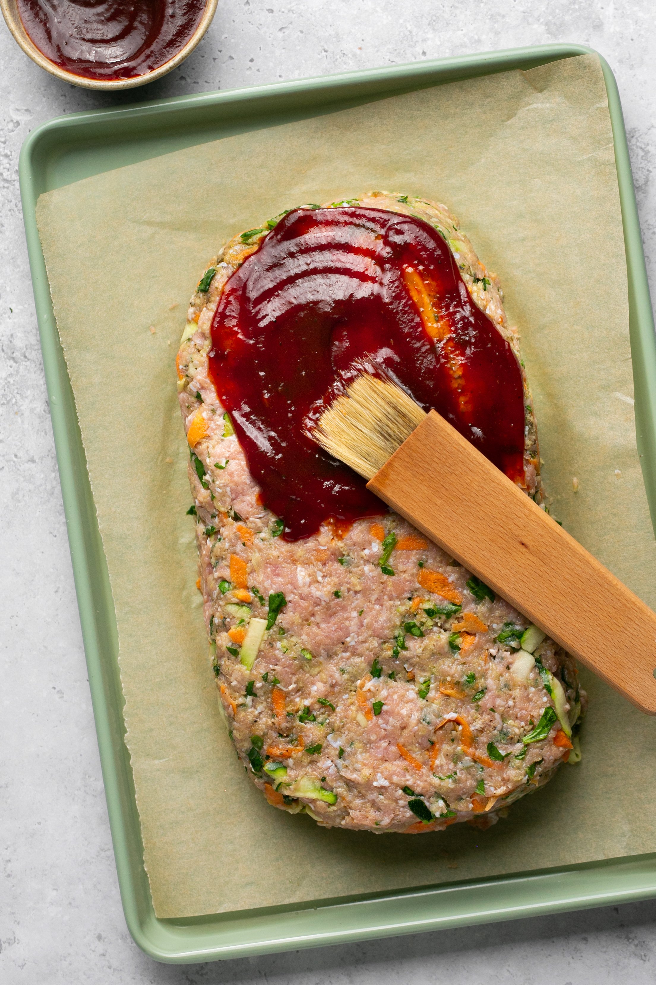 Raw turkey meatloaf mixture shaped into a rectangular loaf sitting on a parchment lined baking sheet on a white counter. Raw meatloaf is being covered with barbecue sauce using a wooden basting brush. A small white bowl of barbecue sauce is set to the side; slightly out of frame.