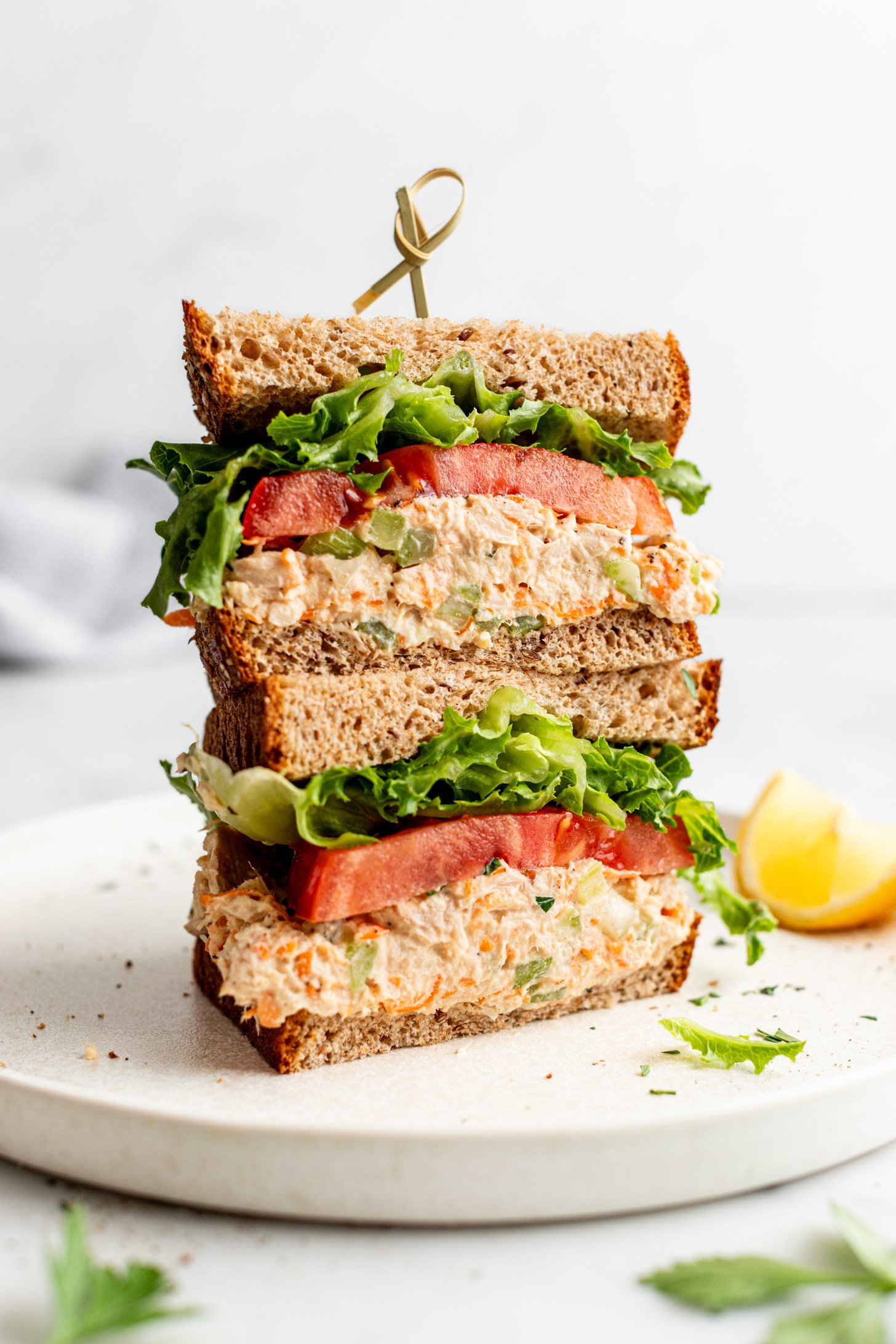 Tuna salad sandwich on wheat bread with lettuce and tomato, cut in half and the halves are stacked on top of one another. There is a wedge of lemon on the plate with the sandwich 