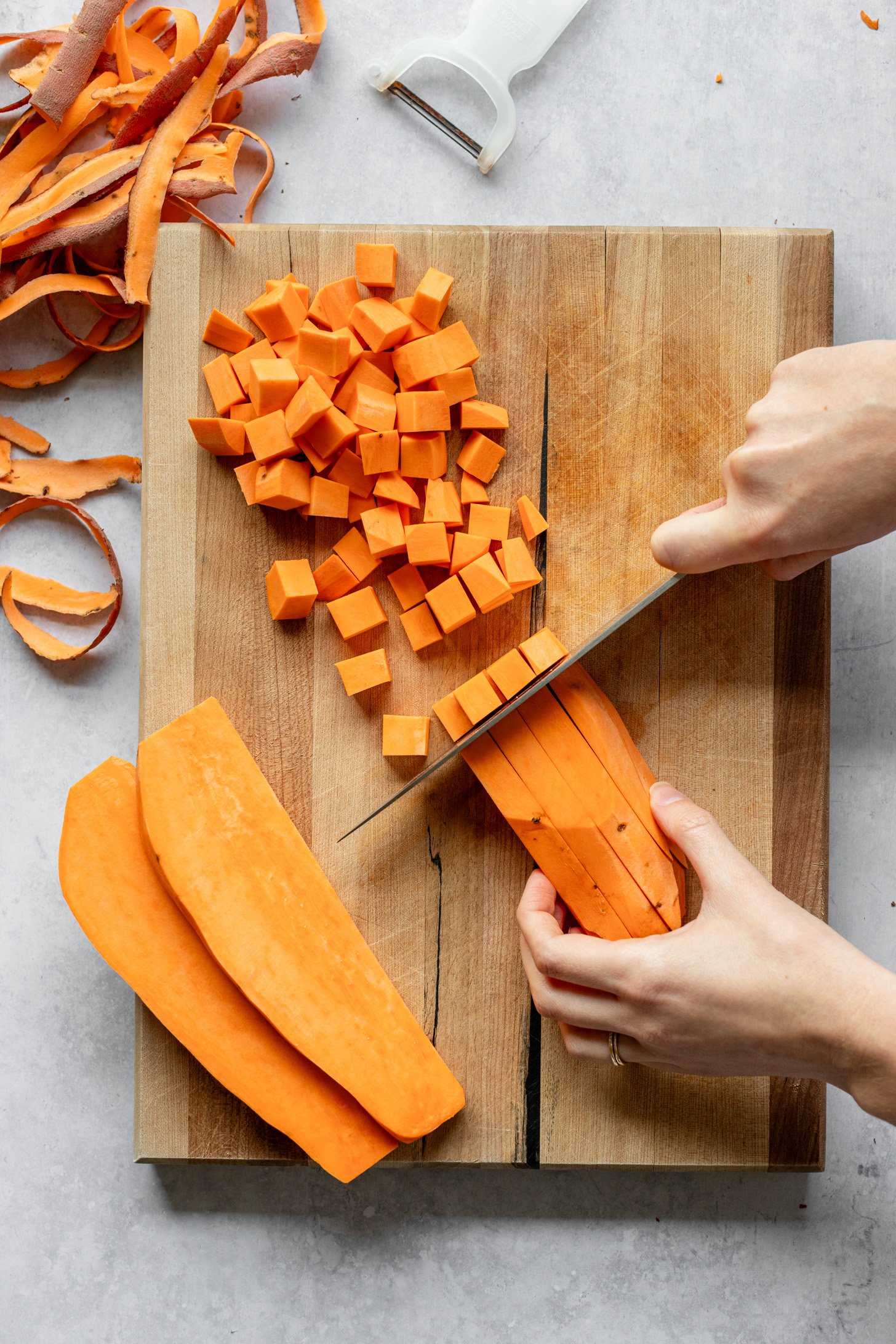 Sweet potato slices are being cut into cubes (diced) on a wooden cutting board on a grey table. Sweet potato skin and a vegetable peeler are off to the side on the table.
