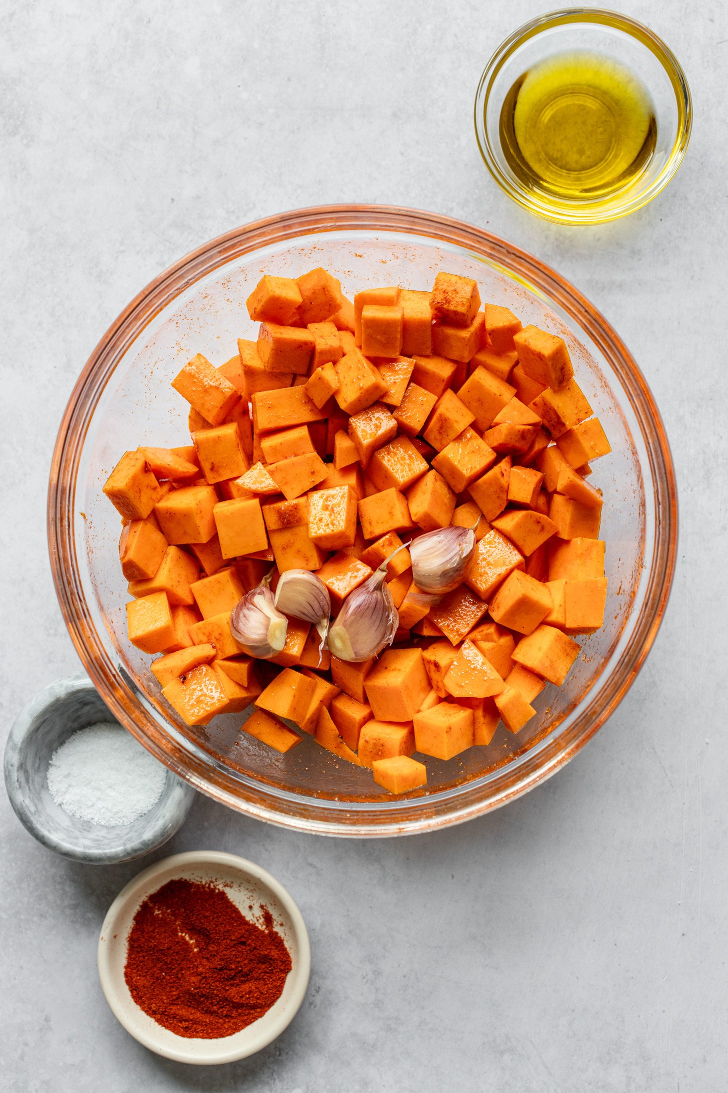 A bowl of diced sweet potato and whole garlic cloves on a grey table There are small glass bowls of olive oil, salt, and spices next to the bowl on the table.