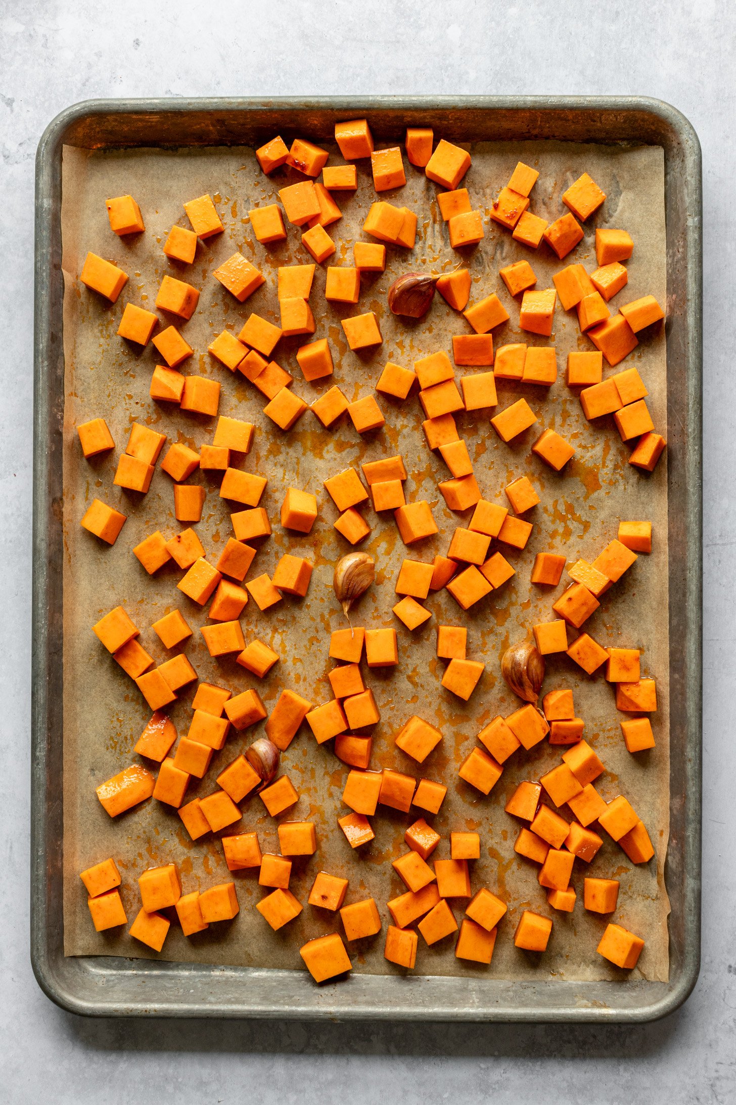 A parchment lined baking sheet with diced sweet potato and garlic cloves on a grey table. 