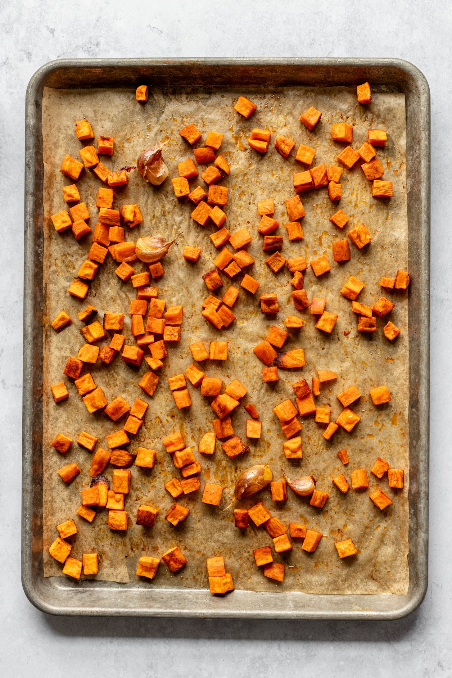 A parchment lined baking sheet with roasted, diced sweet potato and garlic cloves on a grey table. 