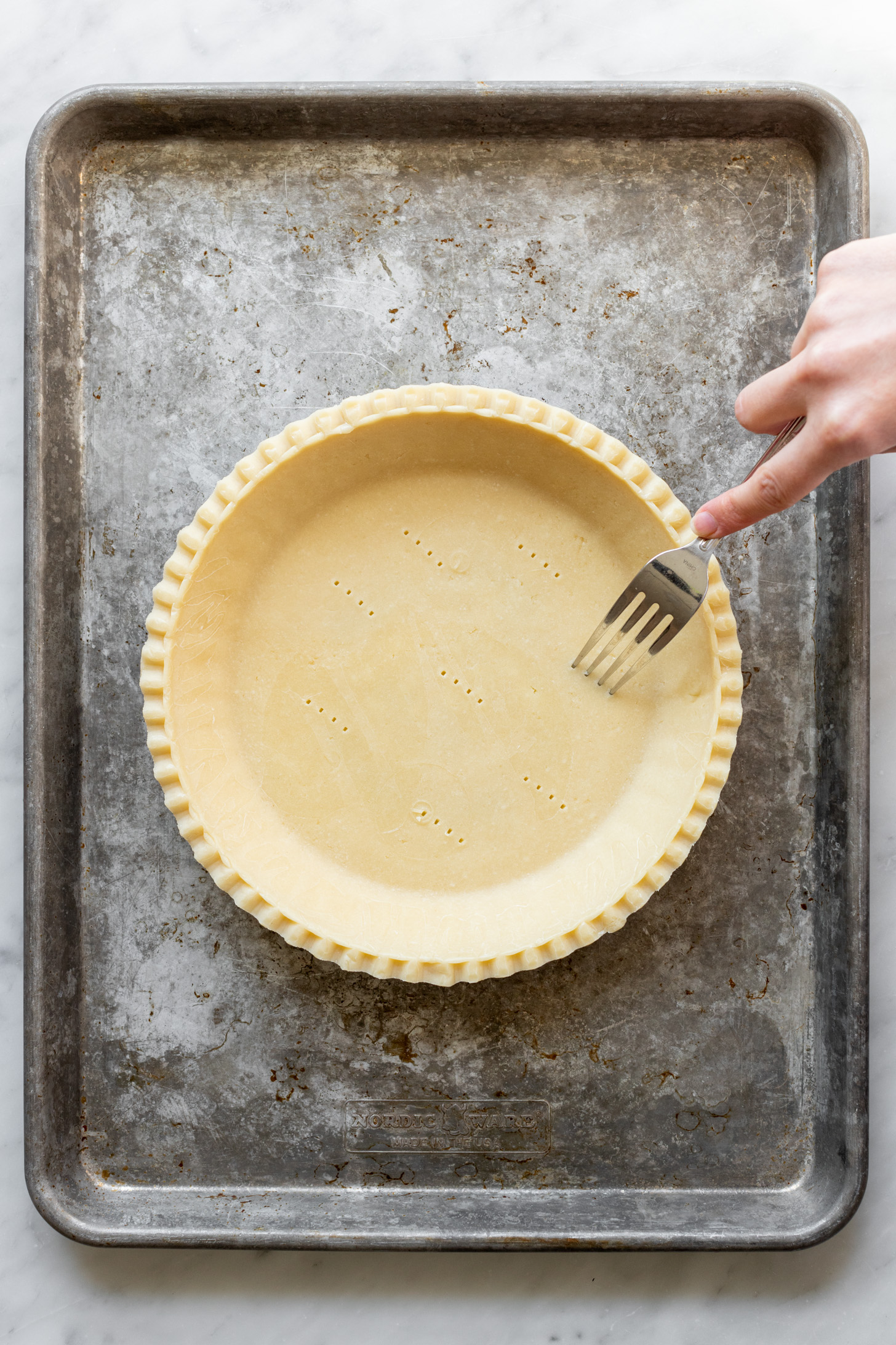 A hand holding a fork pressing tines of the fork into an unbaked pie crust in a pie pan sitting on a sheet pan.