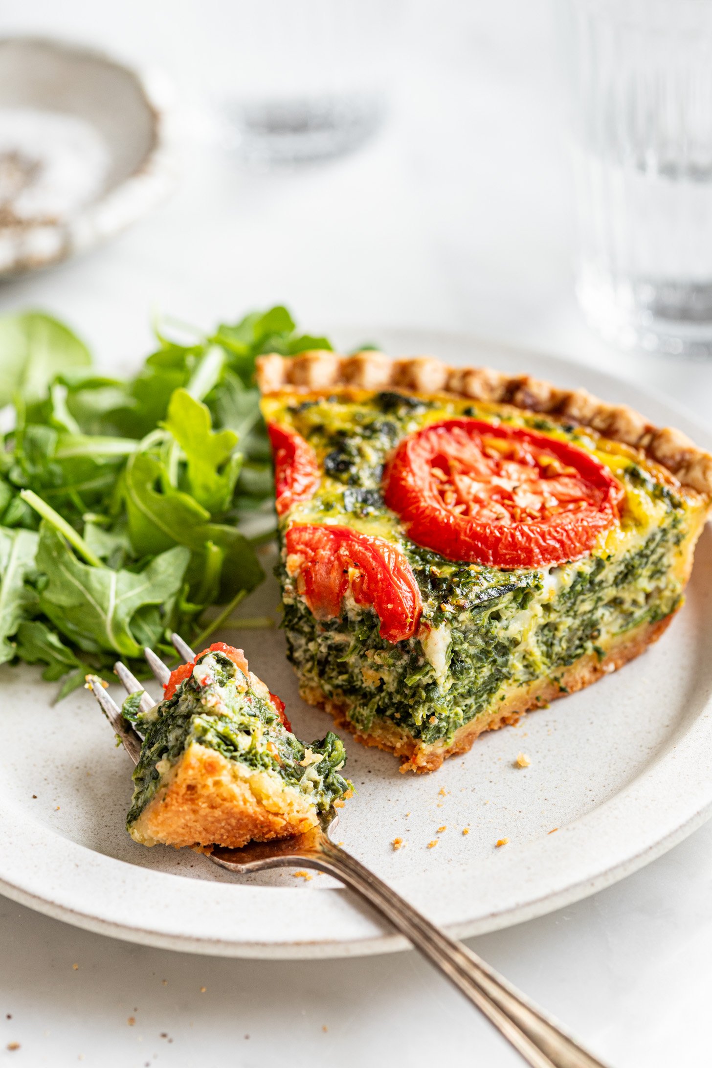 A slice of quiche sitting next to arugula leaves on a white plate. There is a fork holding a piece of quiche also on the plate. Quiche has tomato slices and spinach visible on top. The quiche is sitting on a white marble surface and is surrounded by a few pieces of dishware out of focus in the background. A few crumbs are on the table.
