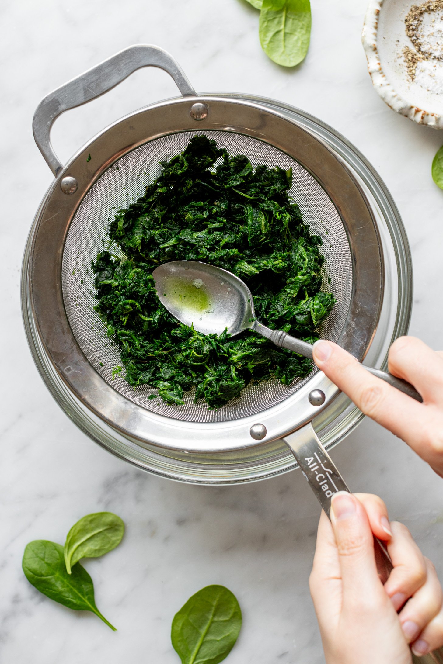 One hand holding a spoon pressing water out of frozen spinach in a strainer. Another hand is holding the strainer which is sitting on top of a glass bowl. Glass bowl is surrounded by a small white bowl filled with salt and pepper. A few baby spinach leaves are strewn on the white marble surface. 