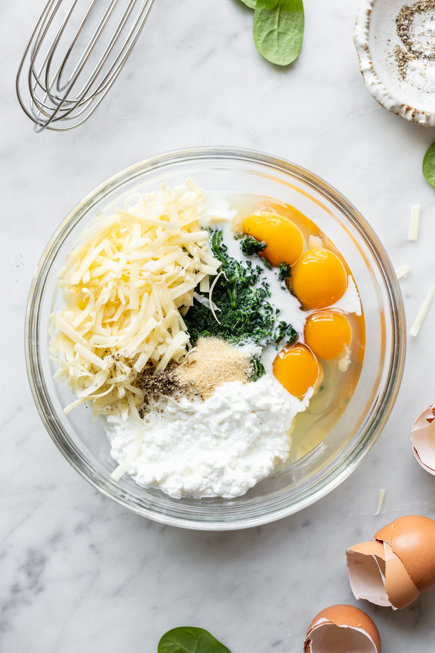 Eggs, spinach, cottage cheese, milk, salt, garlic powder, pepper, and cheddar cheese in a glass bowl sitting on a white marble surface. Strewn about counter are broken eggshells, a few baby spinach leaves, a few cheese shreds, a small bowl with salt and pepper and a whisk.