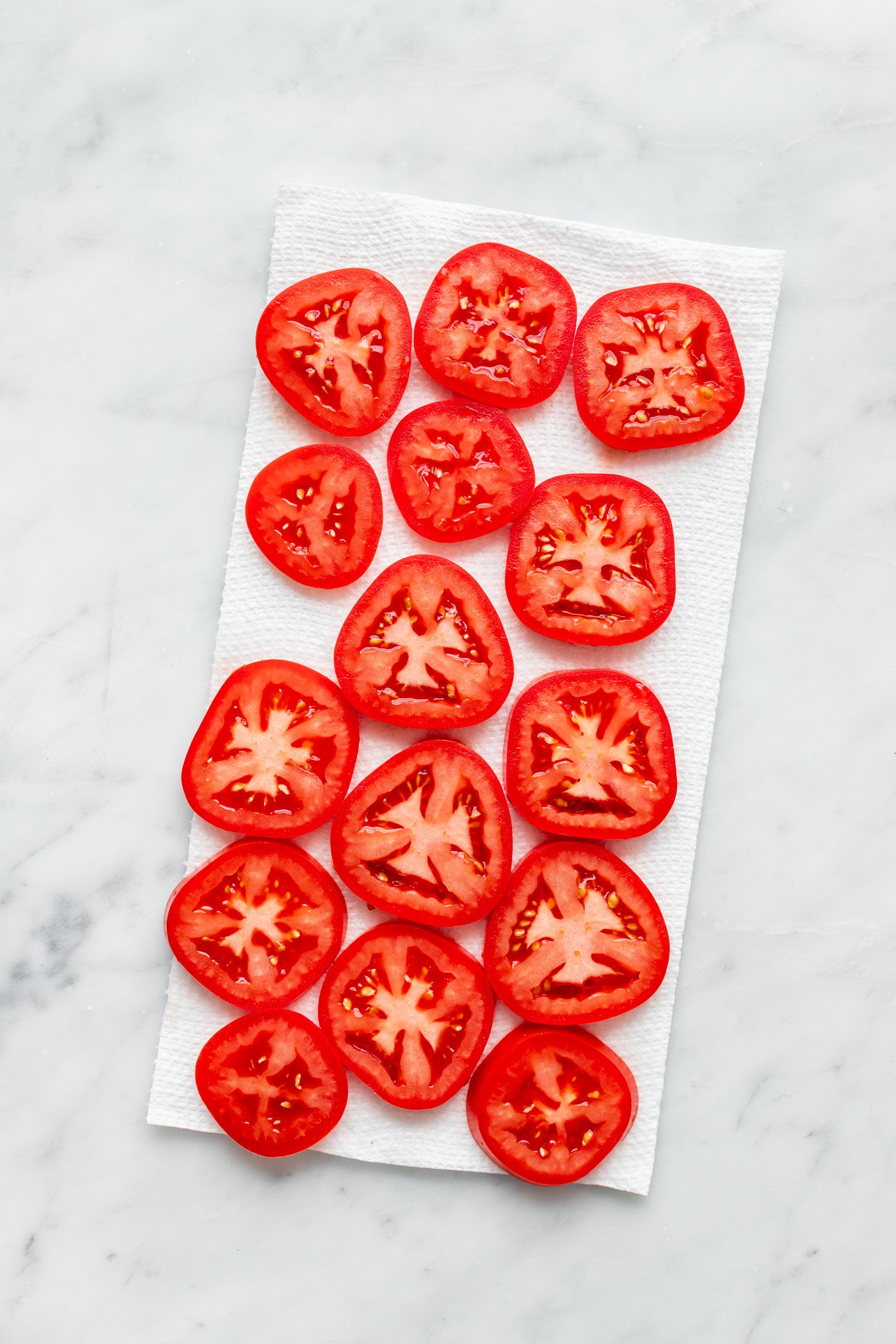Slices of tomato on a paper towel sitting on top of a white marble surface. 