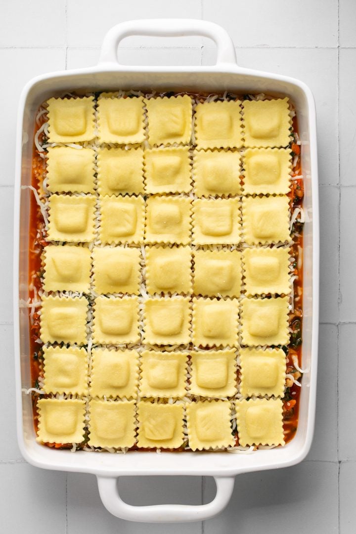 Raviolis arranged in an even layer on top of remaining lasagna layers in a white casserole dish. Casserole dish is sitting on a white surface.