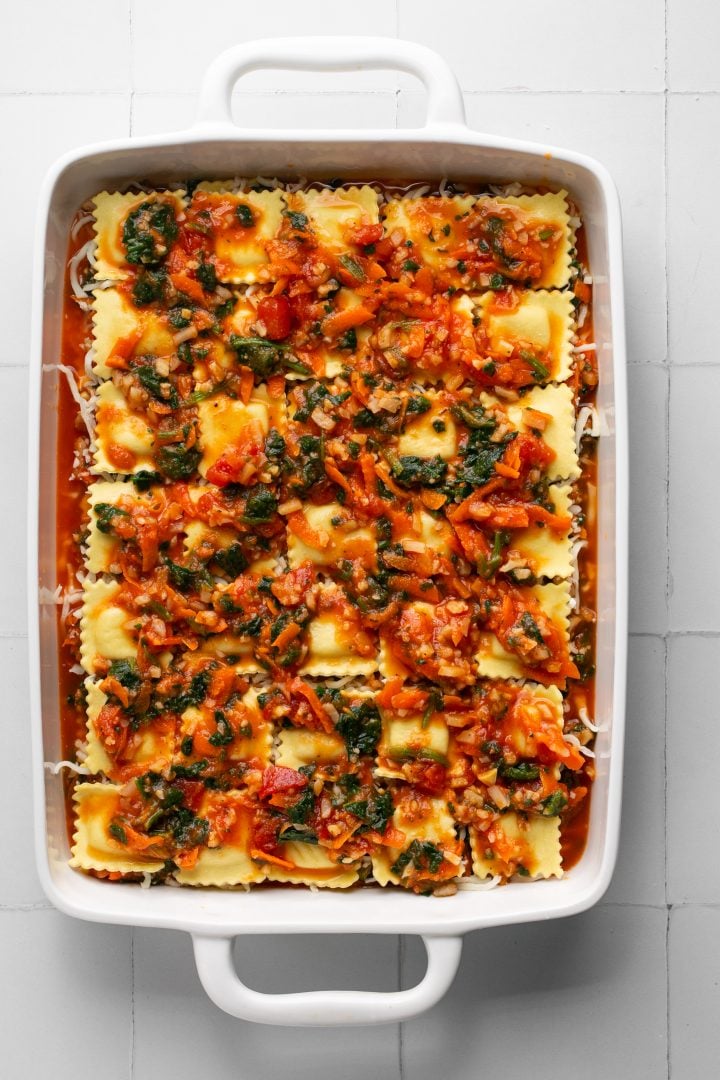 Pasta sauce and vegetable mixture spread evenly on top of a layer of ravioli on top of remaining lasagna layers in a white casserole dish. Casserole dish is sitting on a white surface.
