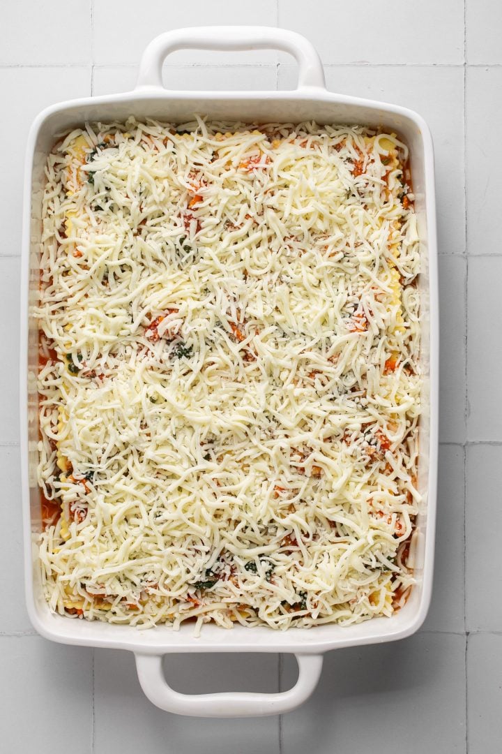 Shredded mozzarella cheese is spread evenly on top of a vegetable and pasta sauce mixture and remaining lasagna layers in a white casserole dish. Casserole dish is sitting on a white surface. Lasagna is not yet cooked.
