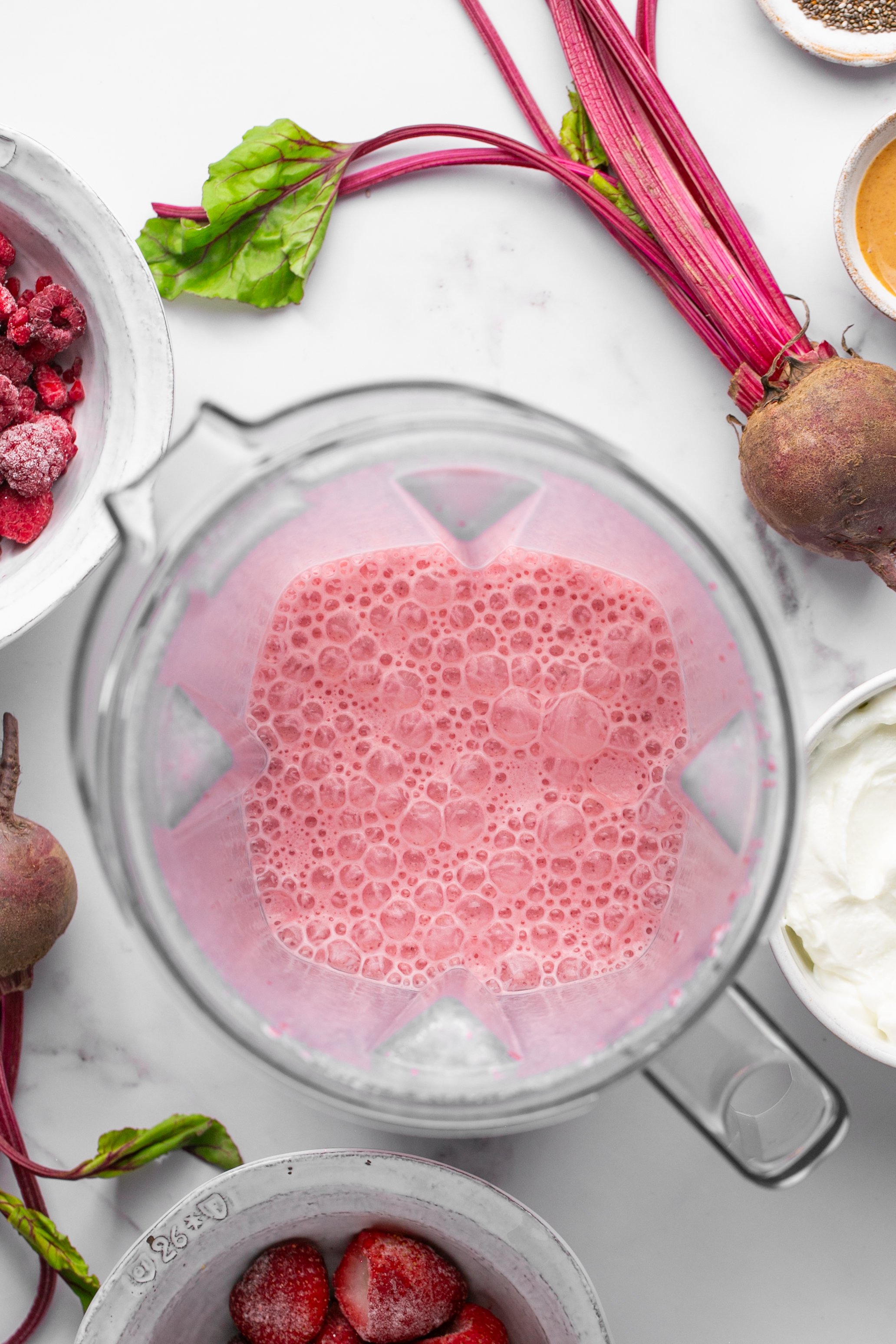 Pureed beet in a blender with milk. Bowls of ingredients for the smoothie are on the table next to the blender.