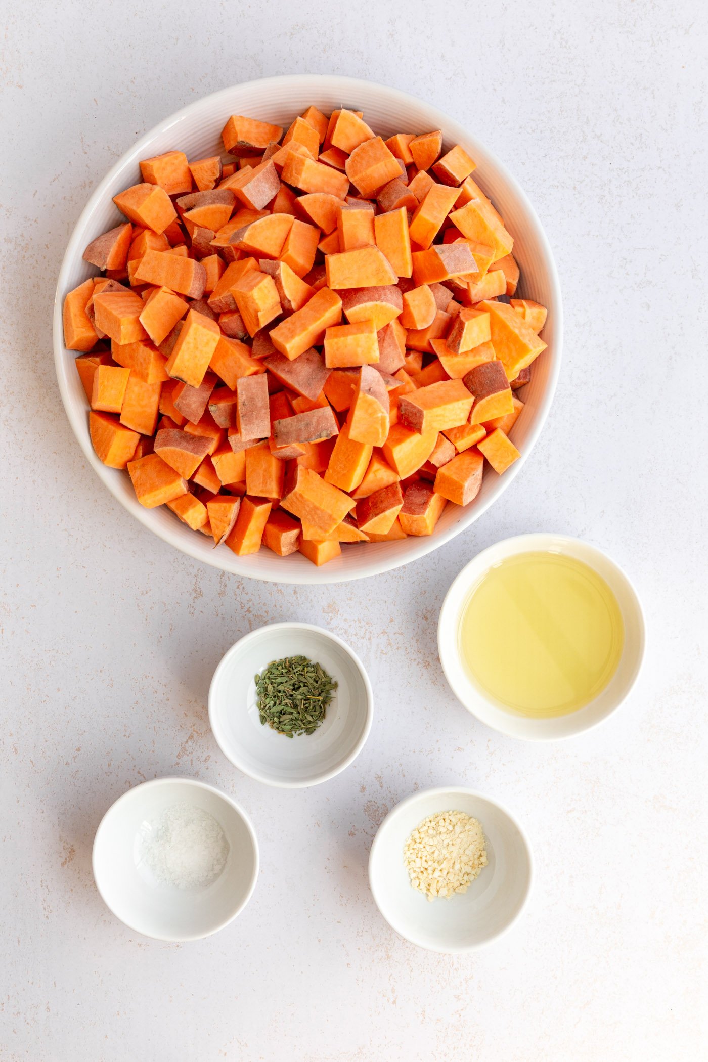 Ingredients for roasted diced sweet potatoes sitting together on a white surface.