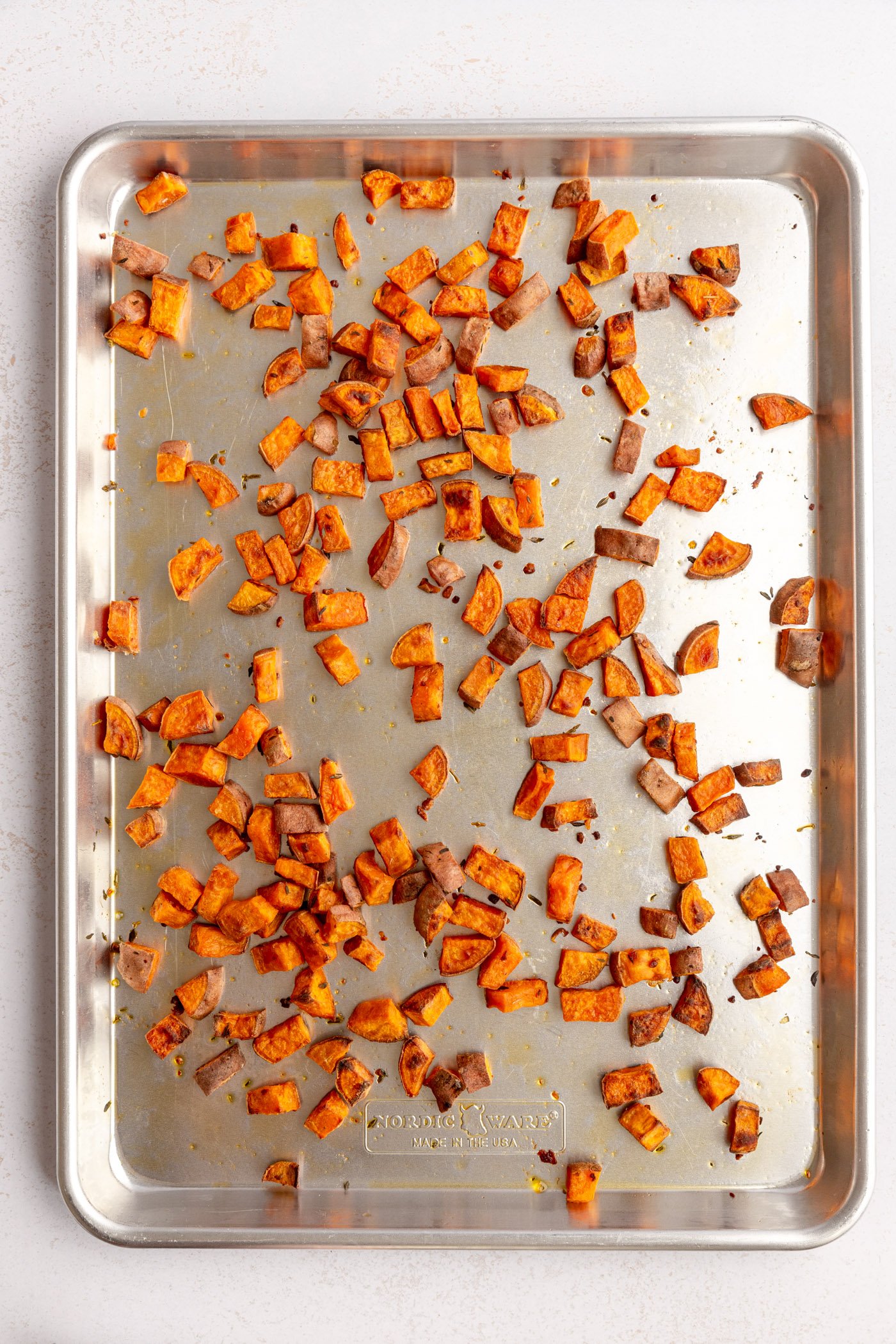 A metal sheet pan filled with evenly spread out roasted diced sweet potatoes. The sheet pan is sitting on a white surface.