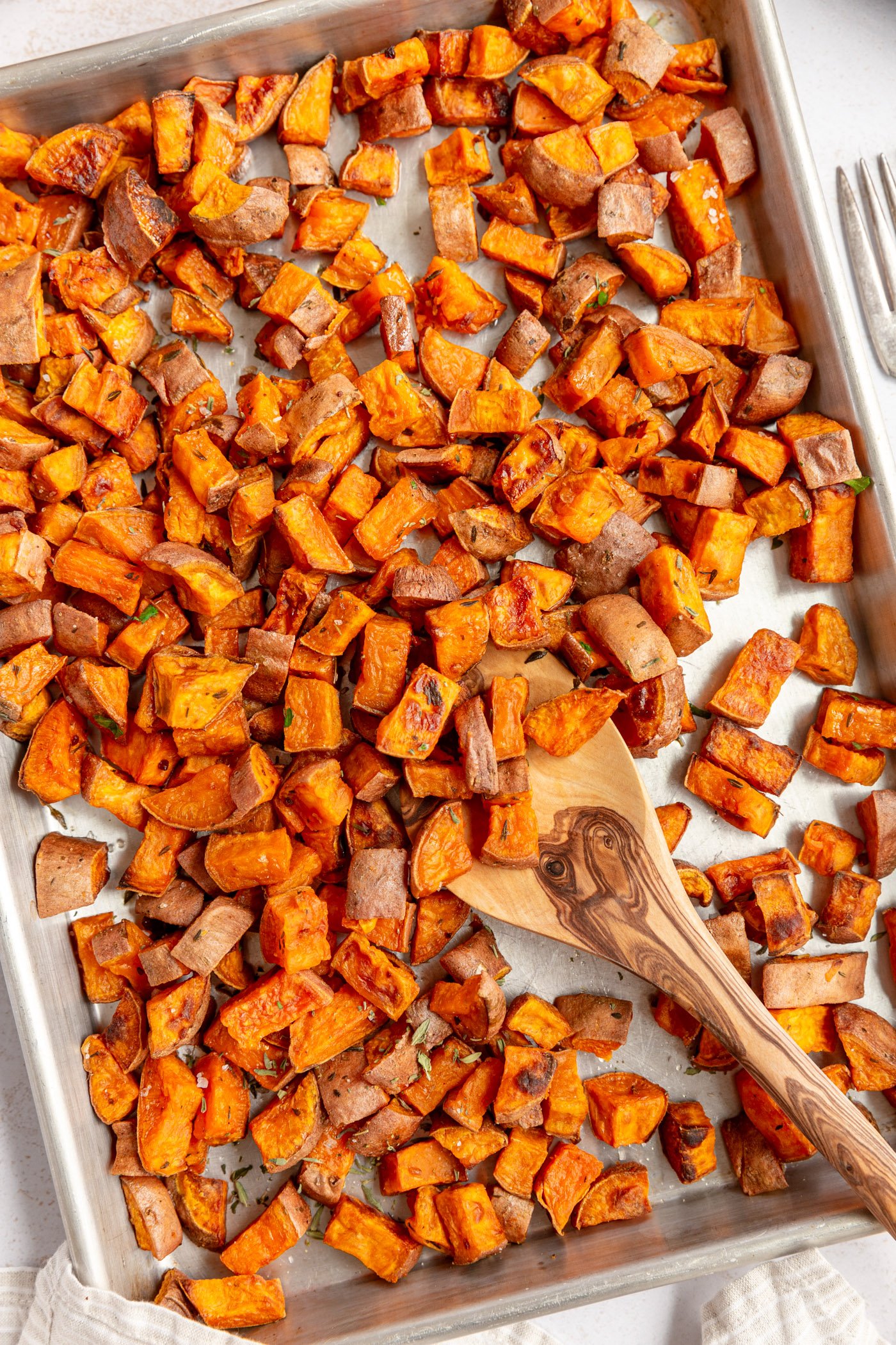 A sheet pan filled with roasted diced sweet potatoes. A wooden spoon is taking taking a helping of the sweet potatoes from the pan. 