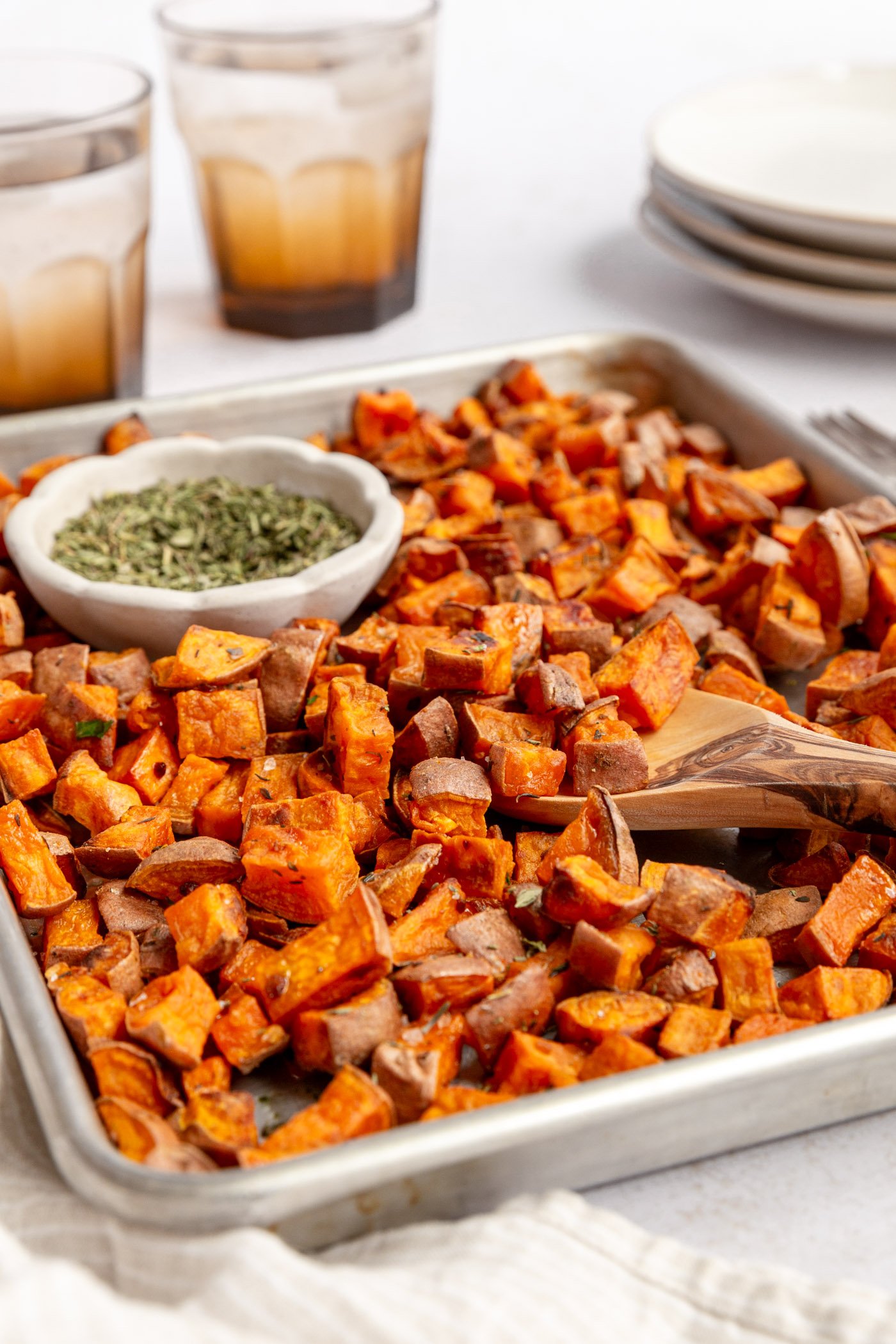 A sheet pan filled with roasted diced sweet potatoes and a small white bowl of dried herbs. A wooden spoon is taking a helping of the sweet potatoes. The sheet pan is surrounded by a napkin, two drinking glasses, and a stack of 3 white plates.