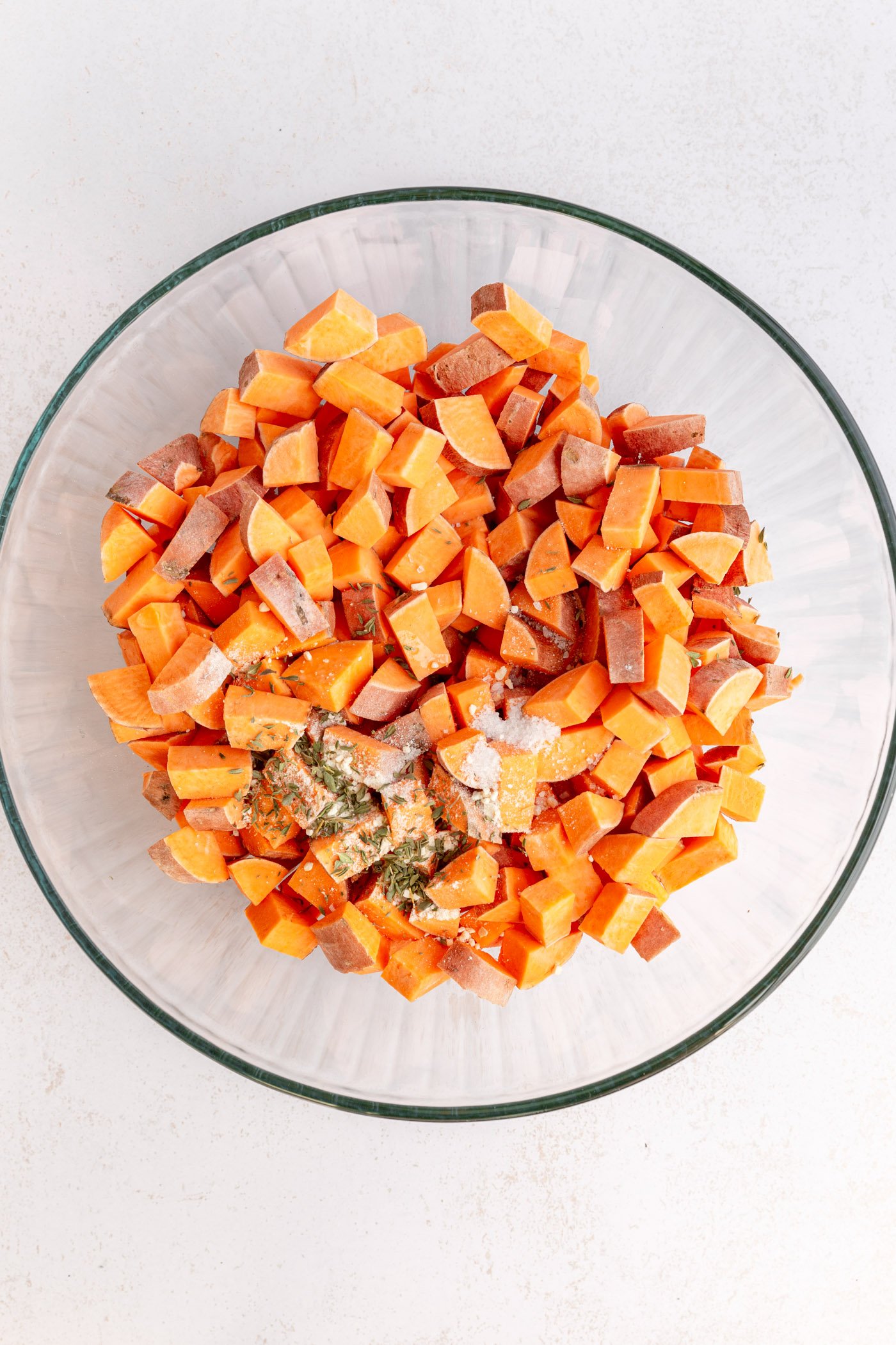 A clear bowl filled with diced sweet potatoes, oil, and seasonings. Bowl is sitting on a white surface.