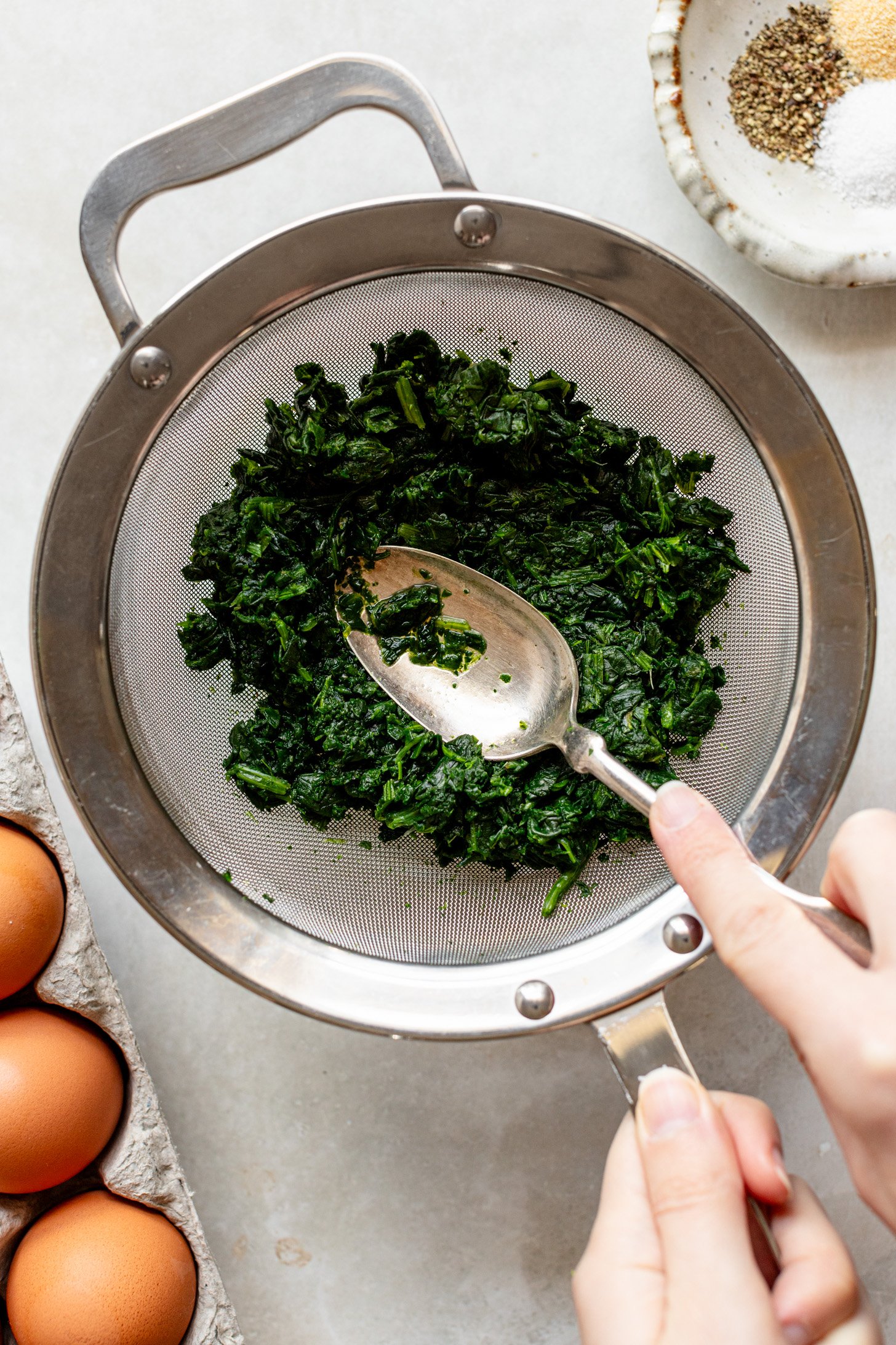 A person draining out thawed frozen spinach over a strainer and using a metal spoon to push all the water out. There is a bowl of seasoning and eggs in the corners of the image.