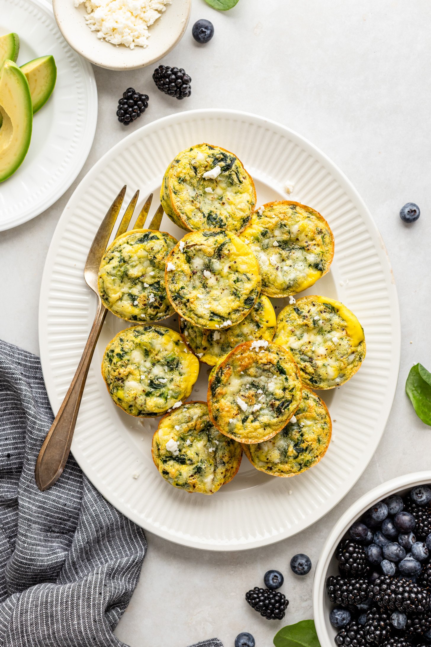 spinach & feta eggs cups on a white platter with a serving fork. There are berries in a bowl and a plate of sliced avocado next to the platter and a gray striped napkin.