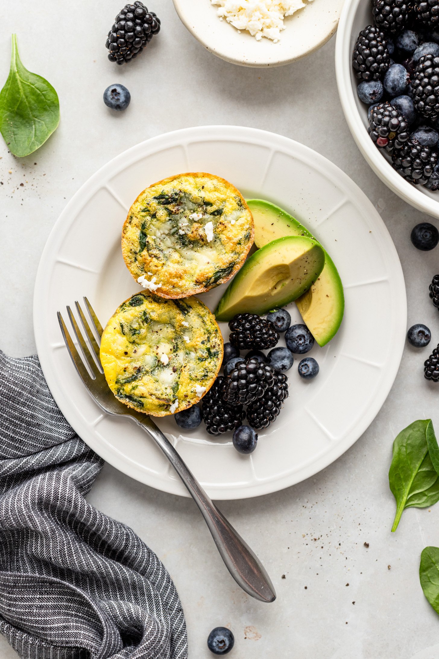 Two spinach feta egg cups on a white plate with avocado and berries. there is a fork on the plate and there is a napkin, spinach leaves, and berries next to it in a bowl