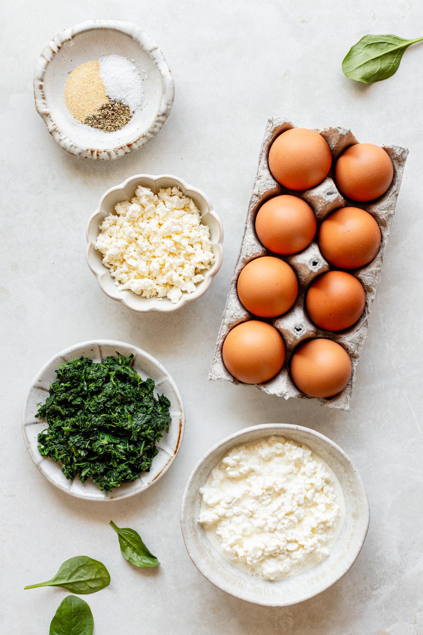 Ingredients for spinach feta egg cups on a white table in bowls and containers before being mixed together.