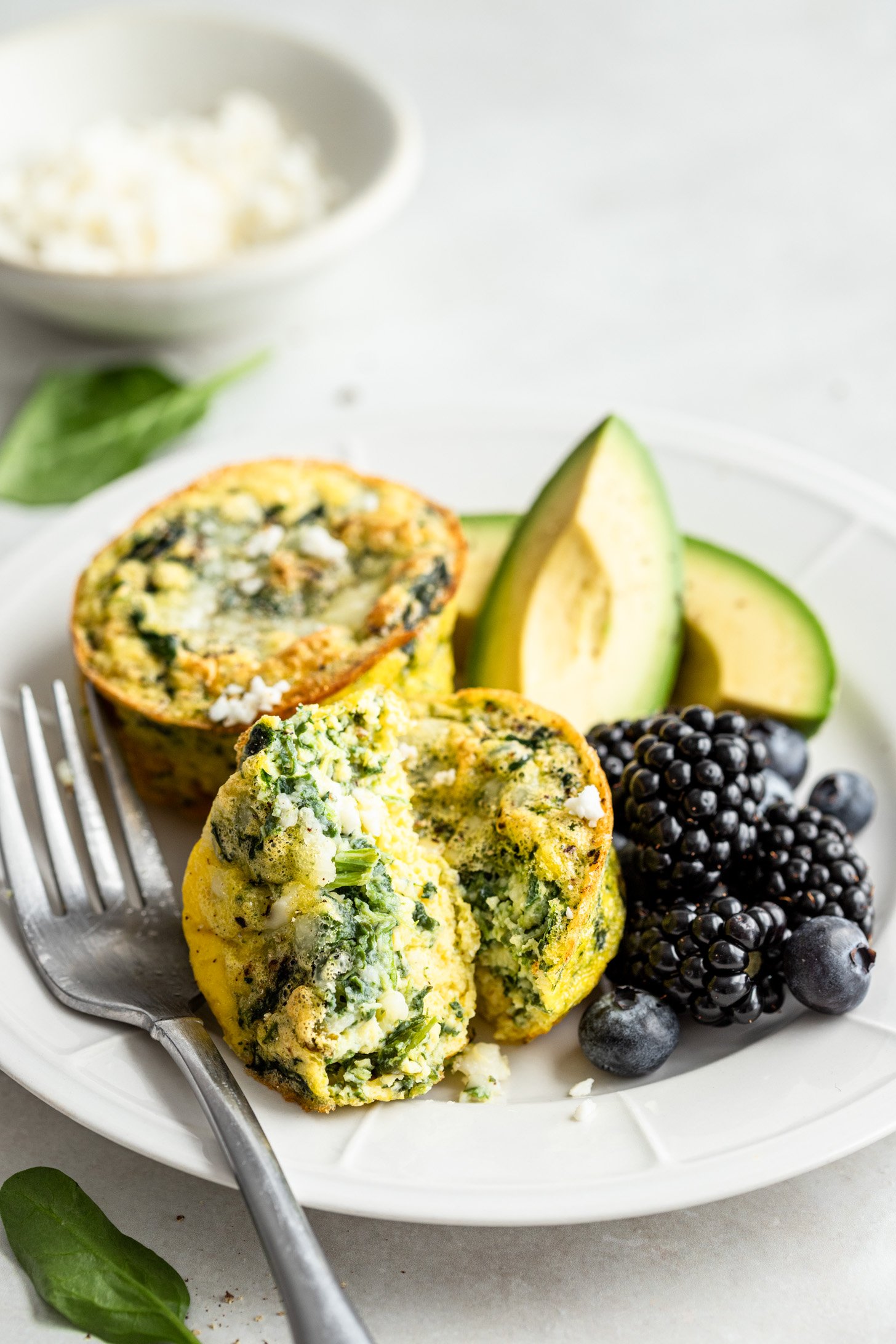 Two feta spinach eggs cups on a plate with avocado and berries. There is a fork on the plate and one egg muffin has been cut in half.