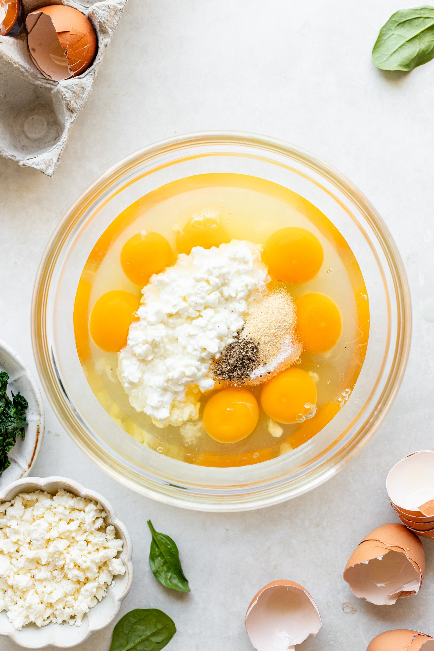 Eggs, cottage cheese, salt, pepper, and garlic powder in a glass bowl. There are egg shells and spinahc leaves and a bowl of feta on the table next to it. 
