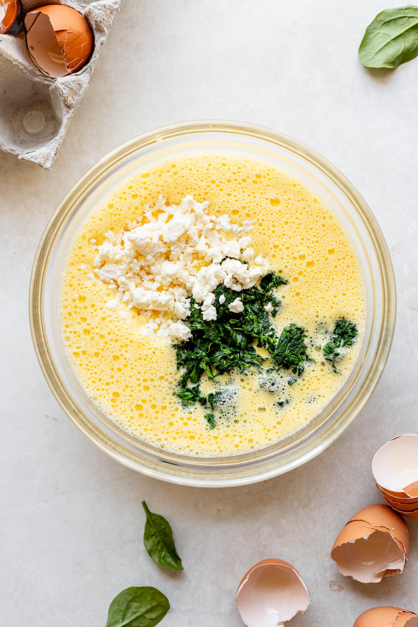 A bowl with pureed egg + cottage cheese mixture. There is feta cheese and spinach on top, but not yet stirred in. There are egg shells and spinach leaves on the table next to the bowl.