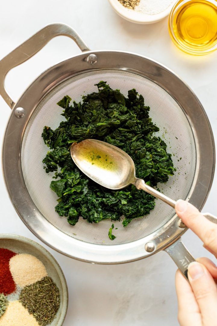 A spoon is being used to push water out of thawed (previously frozen) spinach in a sieve over a glass bowl. Bowl is on a white countertop surrounded by a bowl of spices and a container of oil.