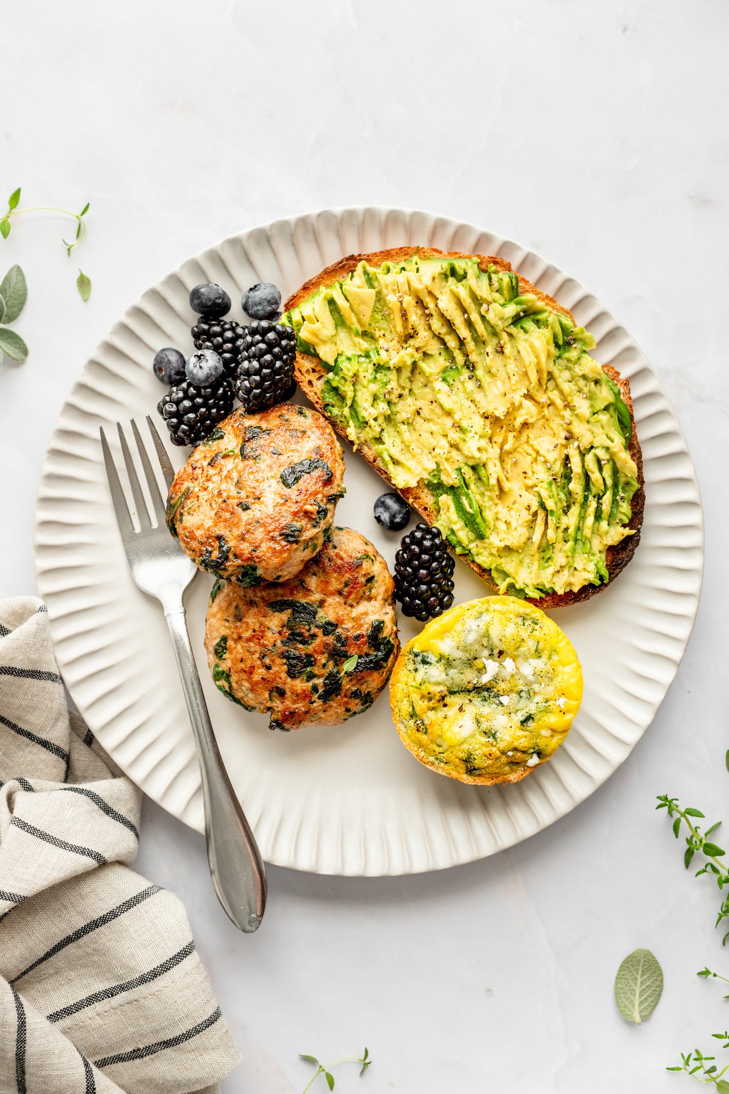 Two spinach & ground turkey breakfast Sausage patties surrounded by an egg "muffin", blackberries, blueberries, and avocado toast, and a fork on a white plate. A striped napkin, sprigs of thyme and sage leaves surround the plate on a white countertop.