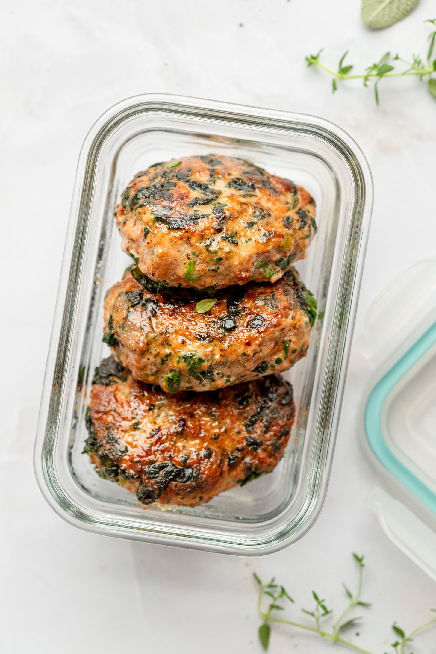 Three cooked Spinach and Ground Turkey Breakfast Sausage patties in a glass food storage container. Food storage container is on a white countertop surrounded by a food storage lid, sprigs of dried thyme, and a dried sage leaf.
