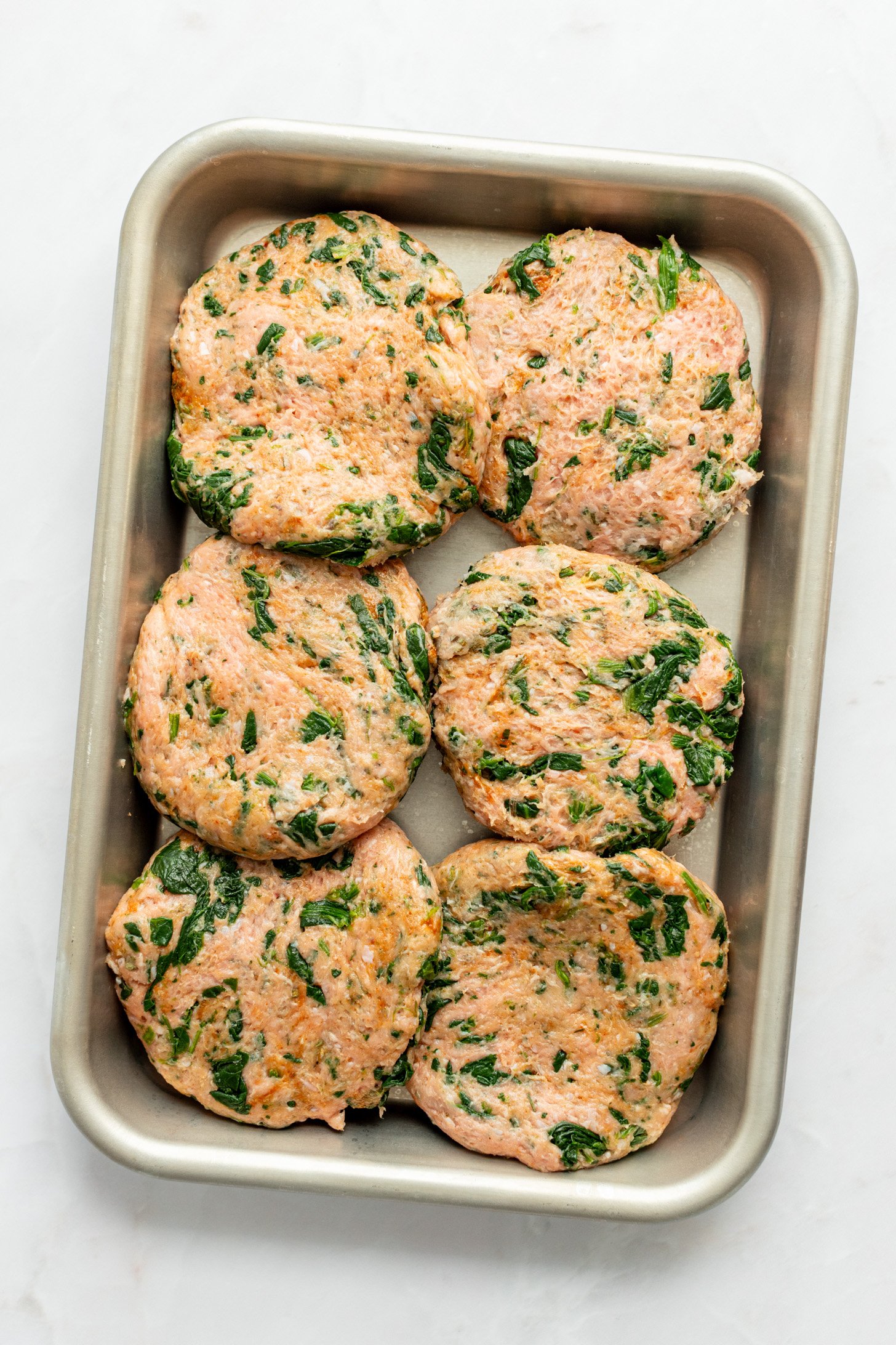 Raw ground turkey mixture for Spinach and Ground Turkey Breakfast Sausages are formed into six patties on a baking sheet sitting on a white countertop.