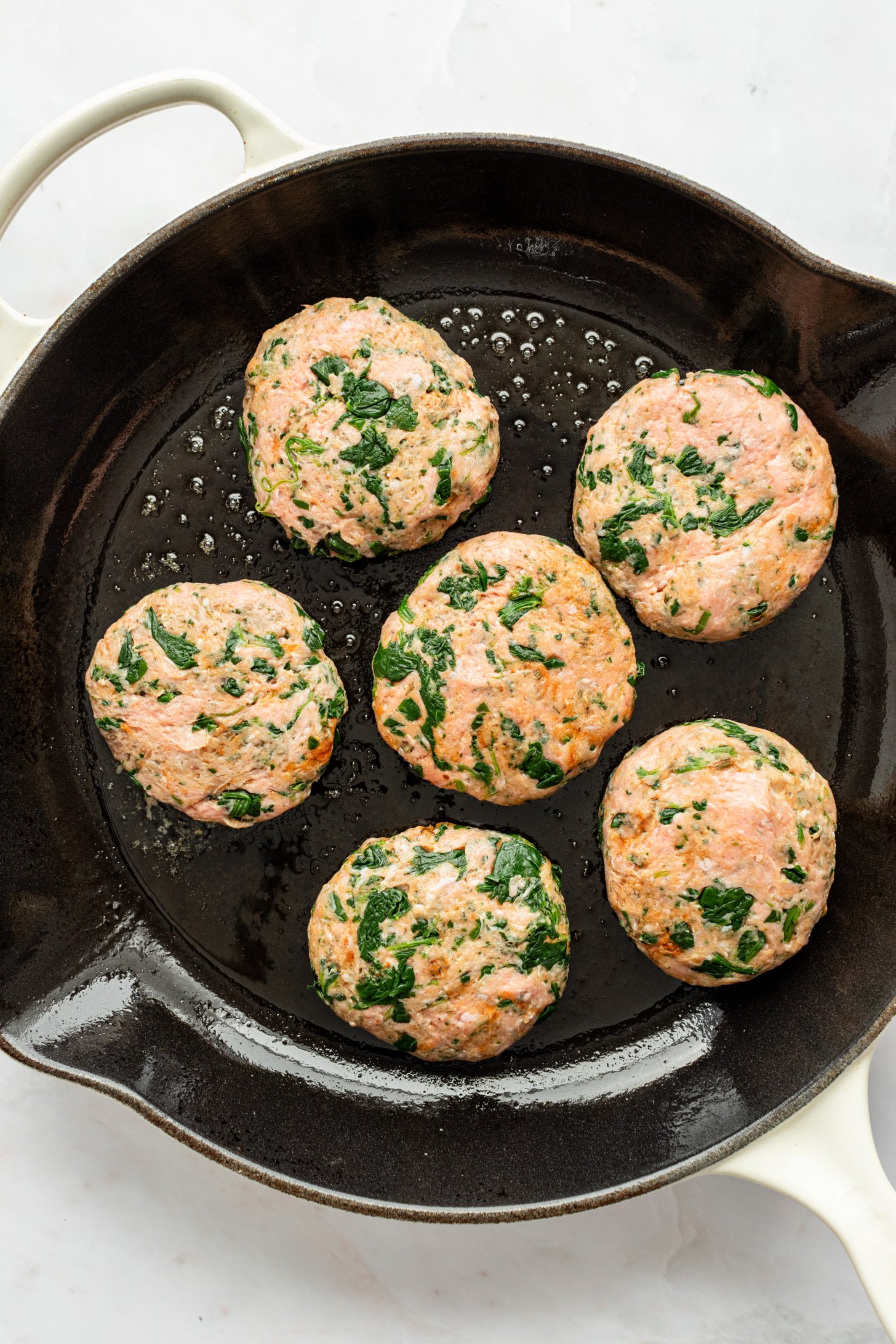 Six raw ground turkey and spinach breakfast sausage patties and oil in a cast iron pan sitting on a white countertop.