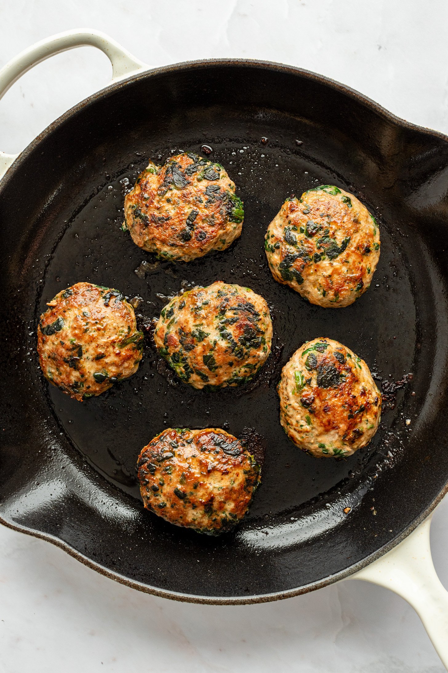 Six cooked ground turkey and spinach breakfast sausage patties and oil in a cast iron pan sitting on a white countertop.
