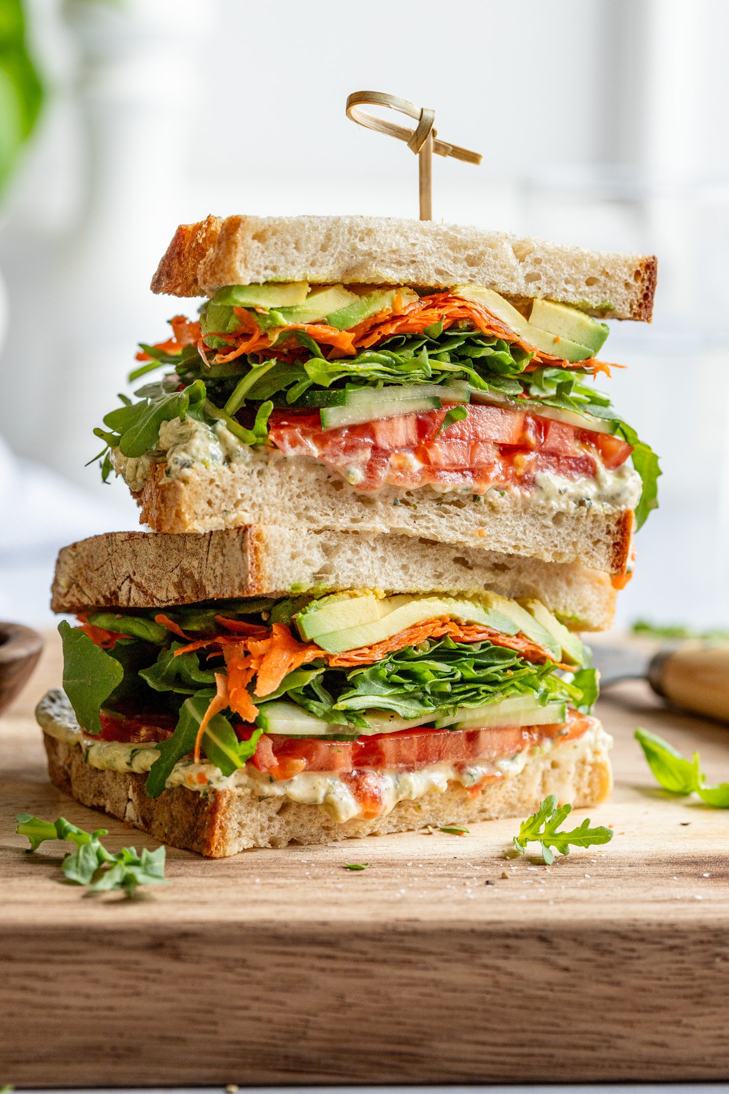 Two halves of a sandwich filled with vegetables and pesto cream cheese stacked on top of each other sitting on a wooden cutting board. There is a wooden food pick speared through the stacked sandwich halves and the sandwiches are surrounded by pieces of greens, and  bowl and knife (out of focus and slightly out of frame)