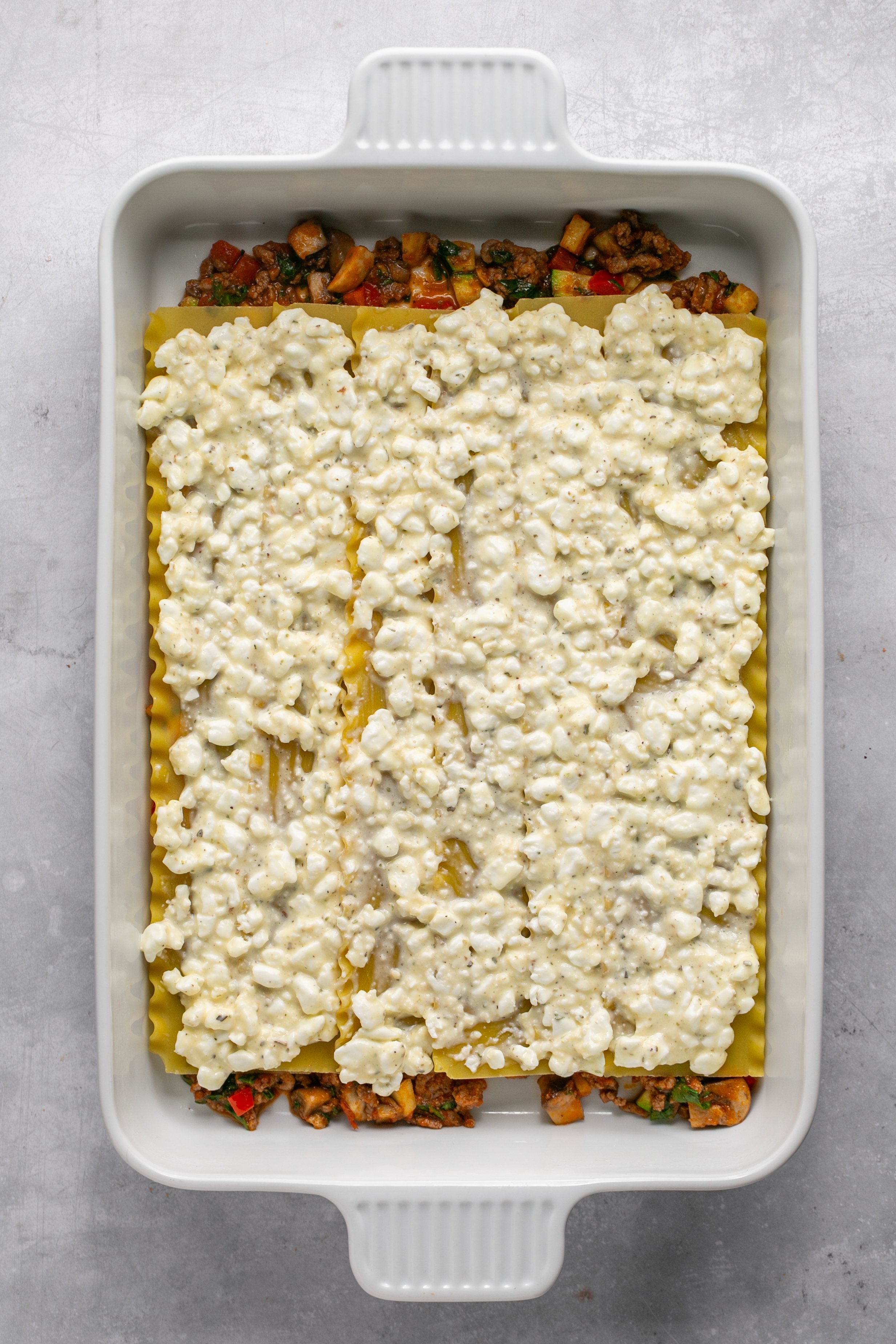 A white casserole dish filled with a layer of vegetable and ground meat mixture, topped with rows of uncooked lasagna noodles. Uncooked lasagna noodles are topped with cheese mixture spread out in an even layer.