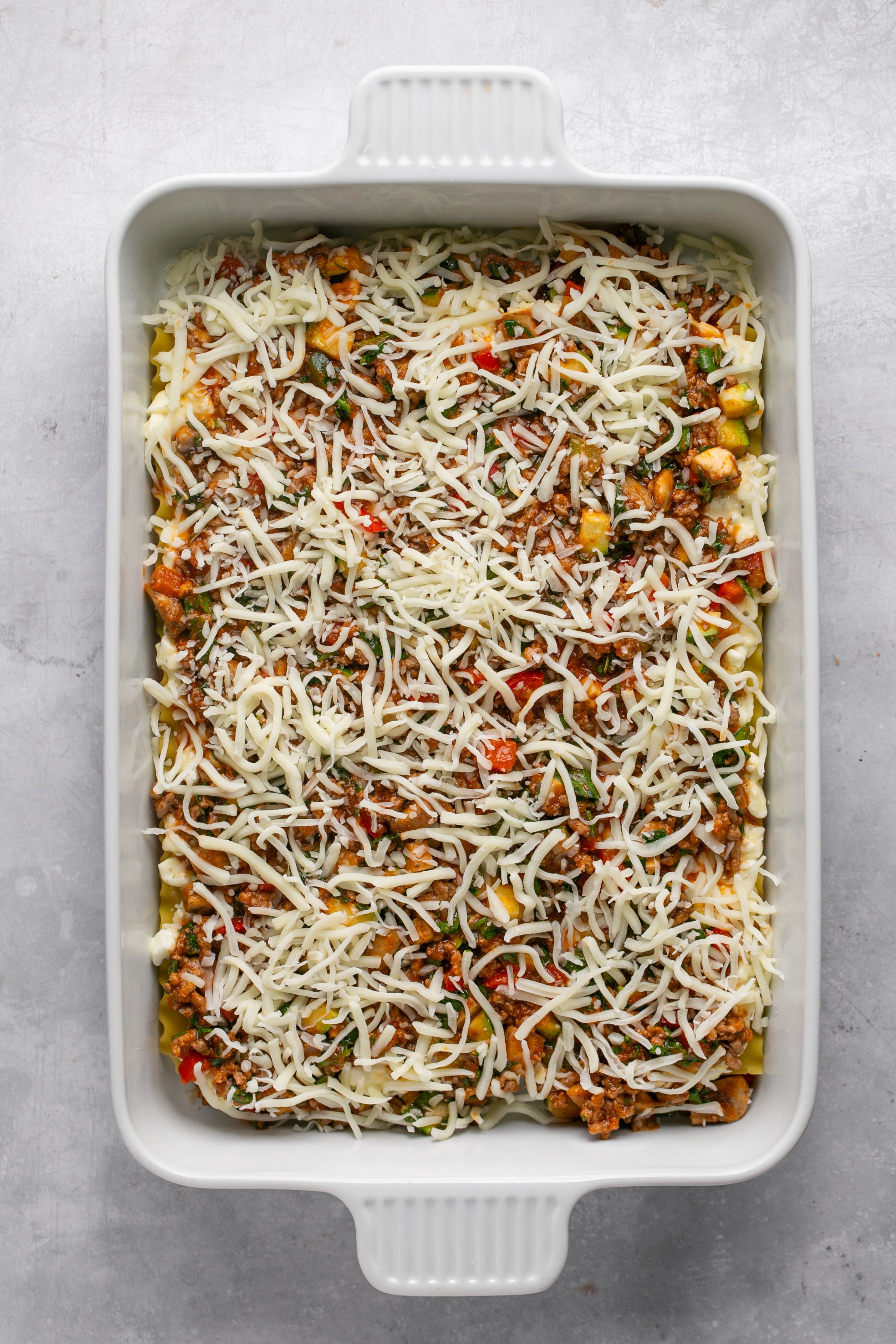 A white casserole dish filled with layers of uncooked lasagna sprinkled with shredded cheese on top.