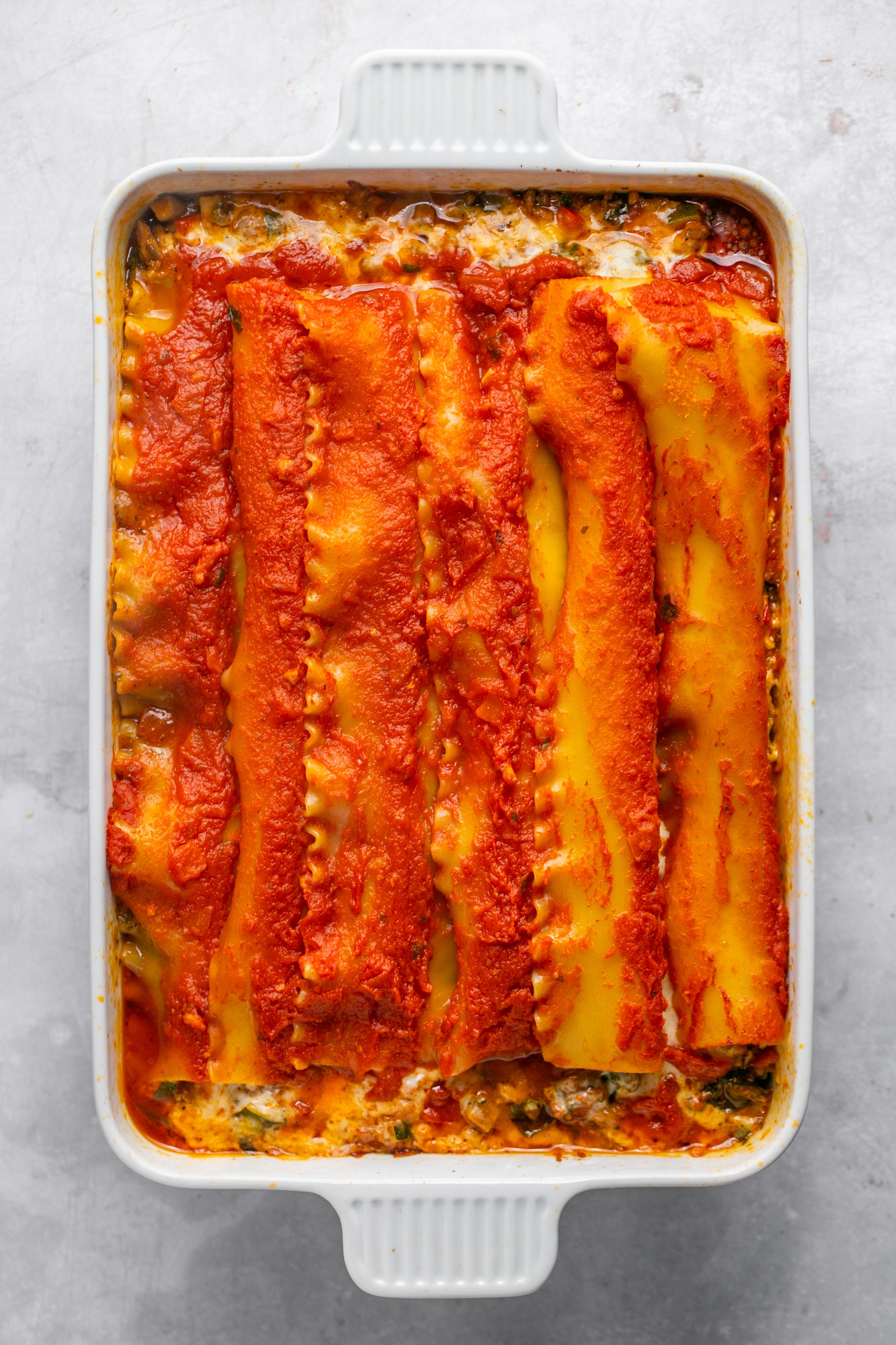 A white casserole dish flled with cooked lasagna sitting on a grey surface. Top layer of the lasagna is noodles and marinara sauce.