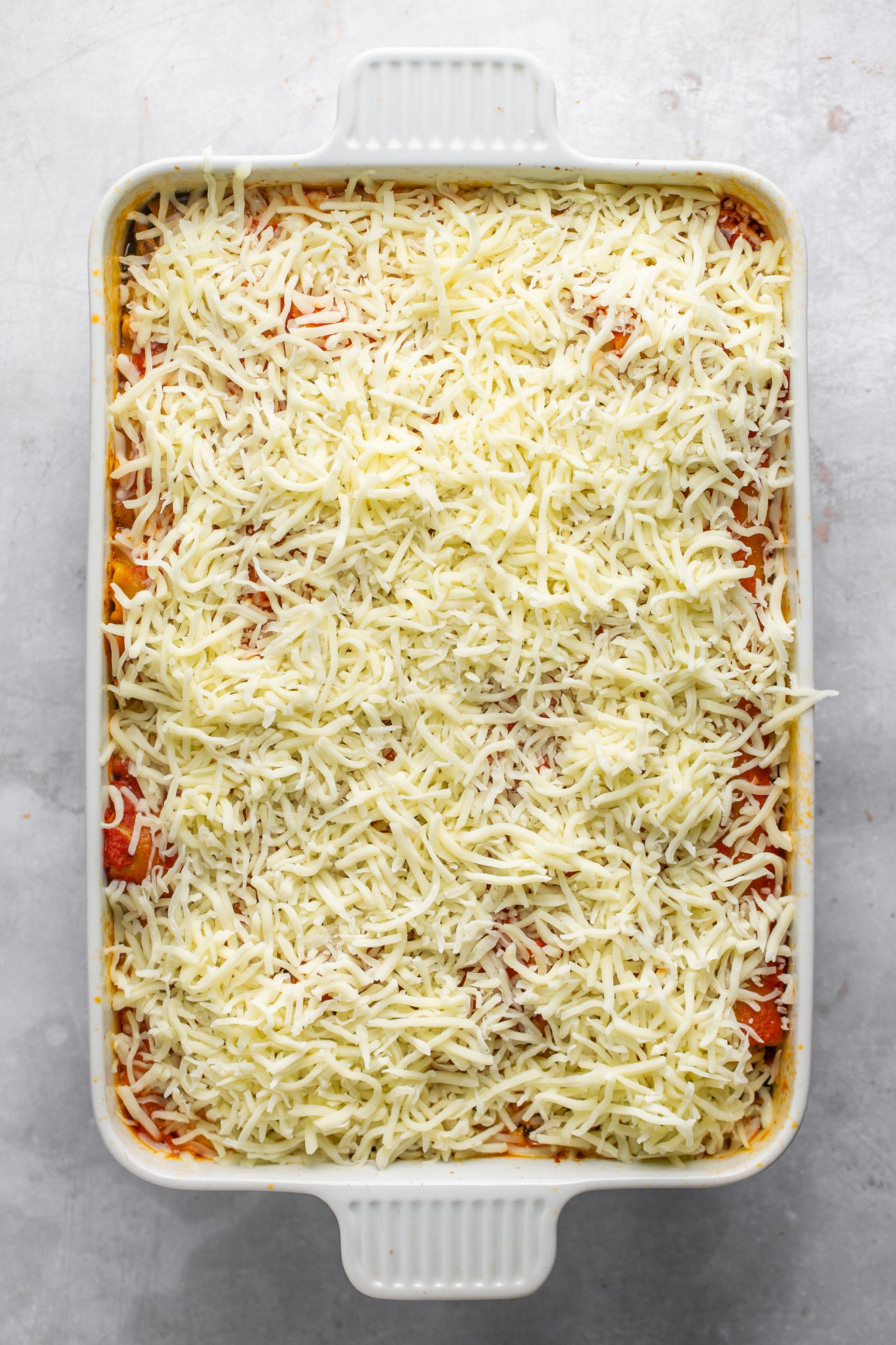 A white casserole dish flled with cooked lasagna topped with unmelted shredded cheese. The casserole dish is sitting on a grey surface.