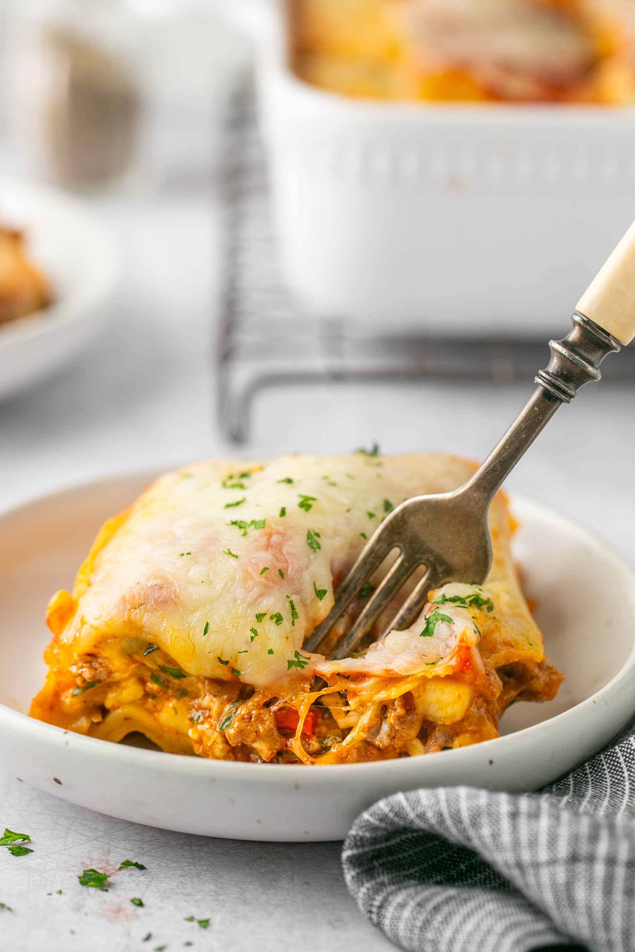 A fork in a slice of lasagna sitting on a white bowl. The bowl is sitting on a grey surface and is surrounded by a napkin. Out of focus in the background is a white casserole dish filled with lasagna on a cooling rack and a few dishes.