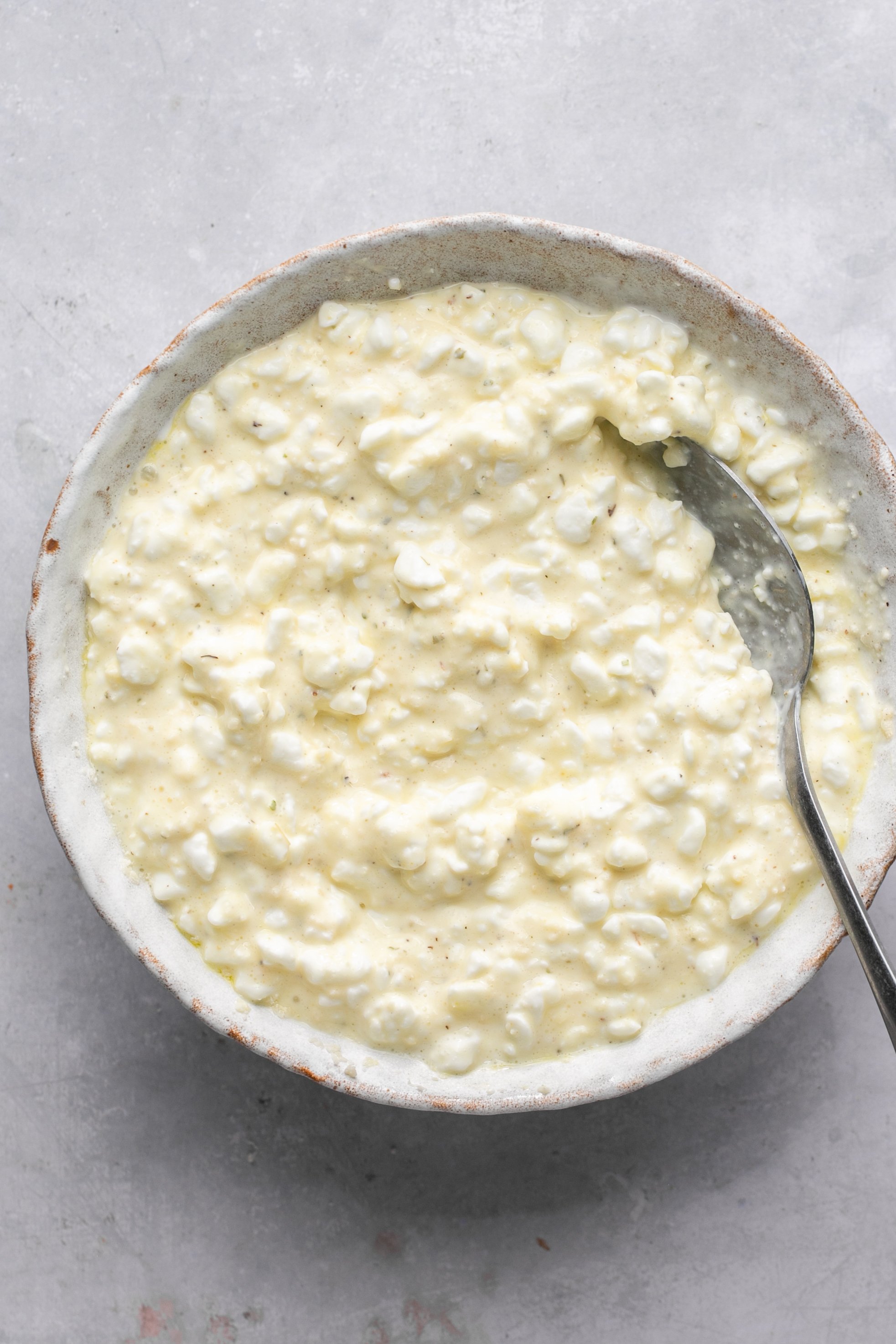 Combined cottage cheese, egg, parmesan cheese, and seasonings in a white bowl sitting on a grey surface. A spoon sitting in the cheese mixture in the bowl.