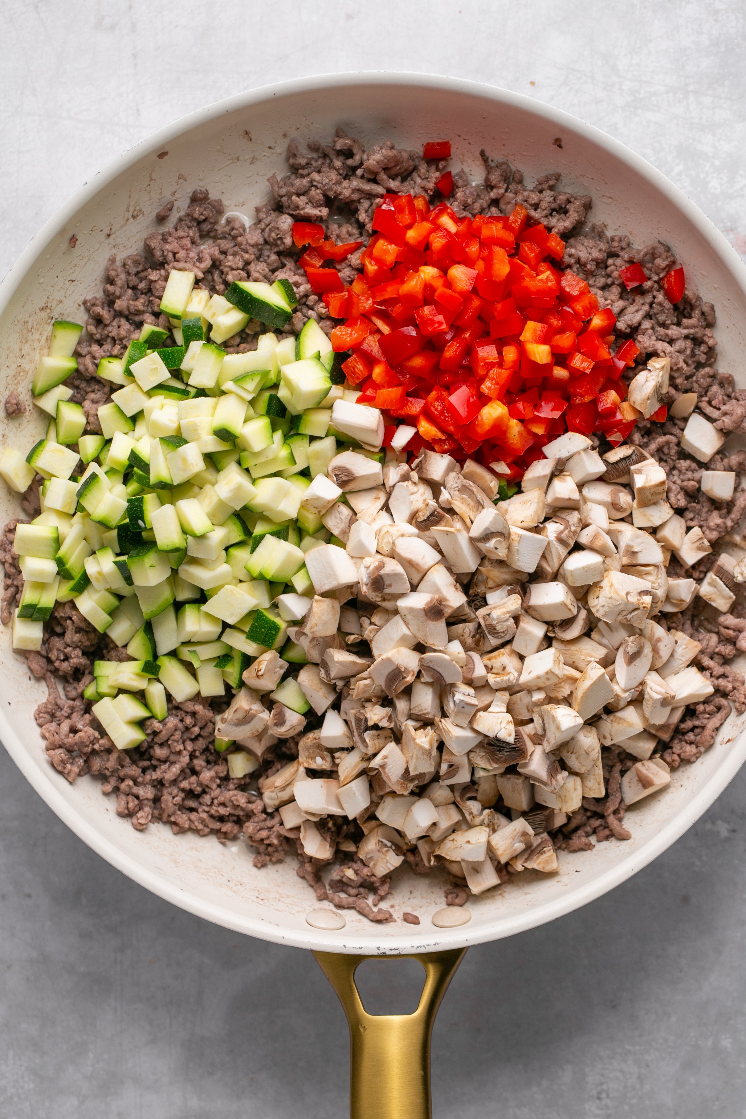 Raw, chopped zucchni, pepper, and mushroom on top of cooked ground meat in a skillet sitting on a grey surface.