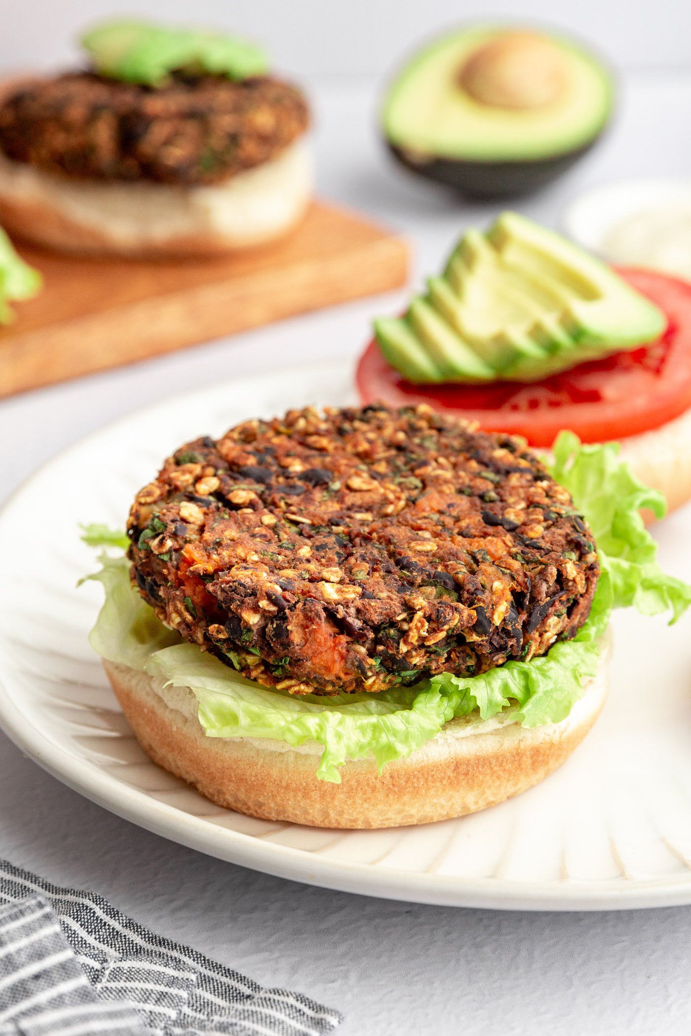 Black bean sweet potato burgers being assembled on burger buns with lettuce, tomato slices, and sliced avocado. They burgers are on a wooden cutting board and there is a striped napkin and small bowl of mayo next to the board.  