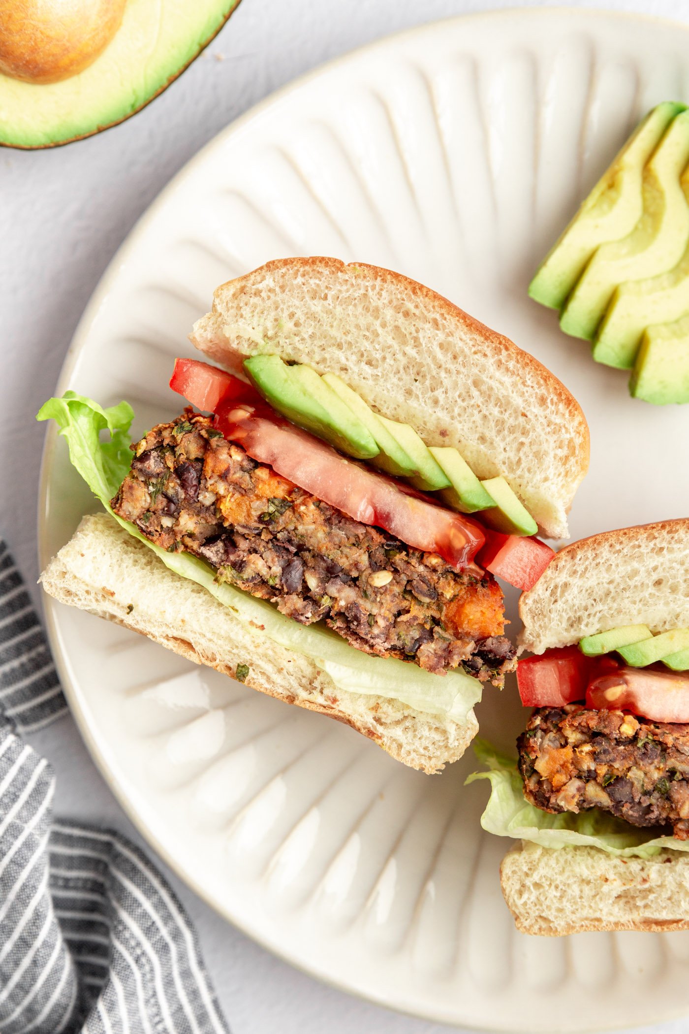 A black bean sweet potato burger on a bun with veggies sliced in half to show the inside texture and turned on its side on a white plate. There is sliced avocado on the plate with the burger.
