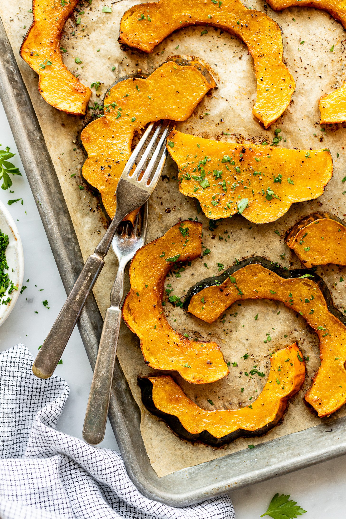 Parchment lined baking sheet with slices of honey roasted acorn squash and two forks. The acorn squash slices are topped with flecks of fresh parsley. The baking sheet is surrounded by a white and blue napkin, a small bowl with chopped parlsey, and some parsley sprinkled on the countertop.