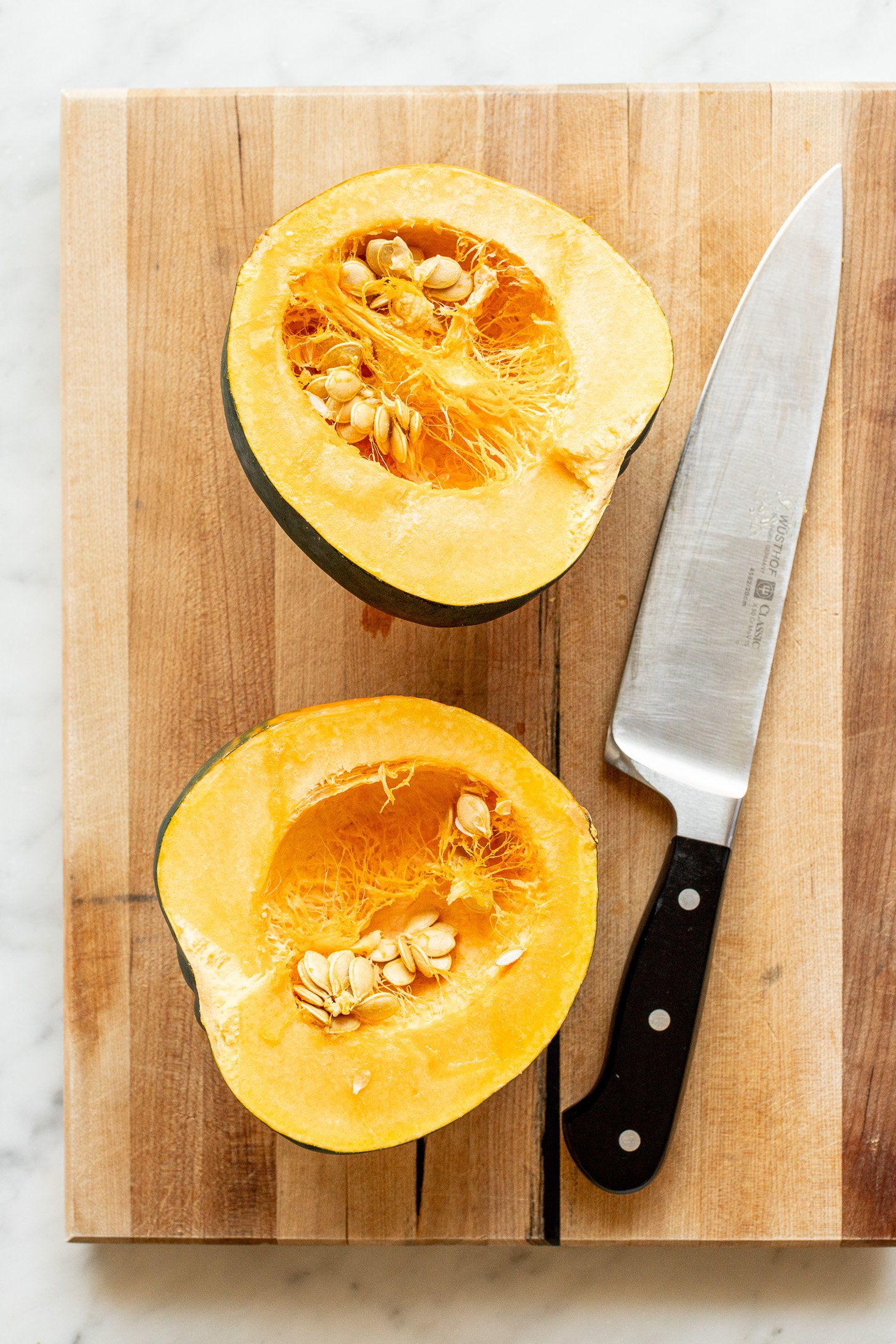 Two halves of an acorn squash along with a chef's knife sitting on a wooden cutting board.