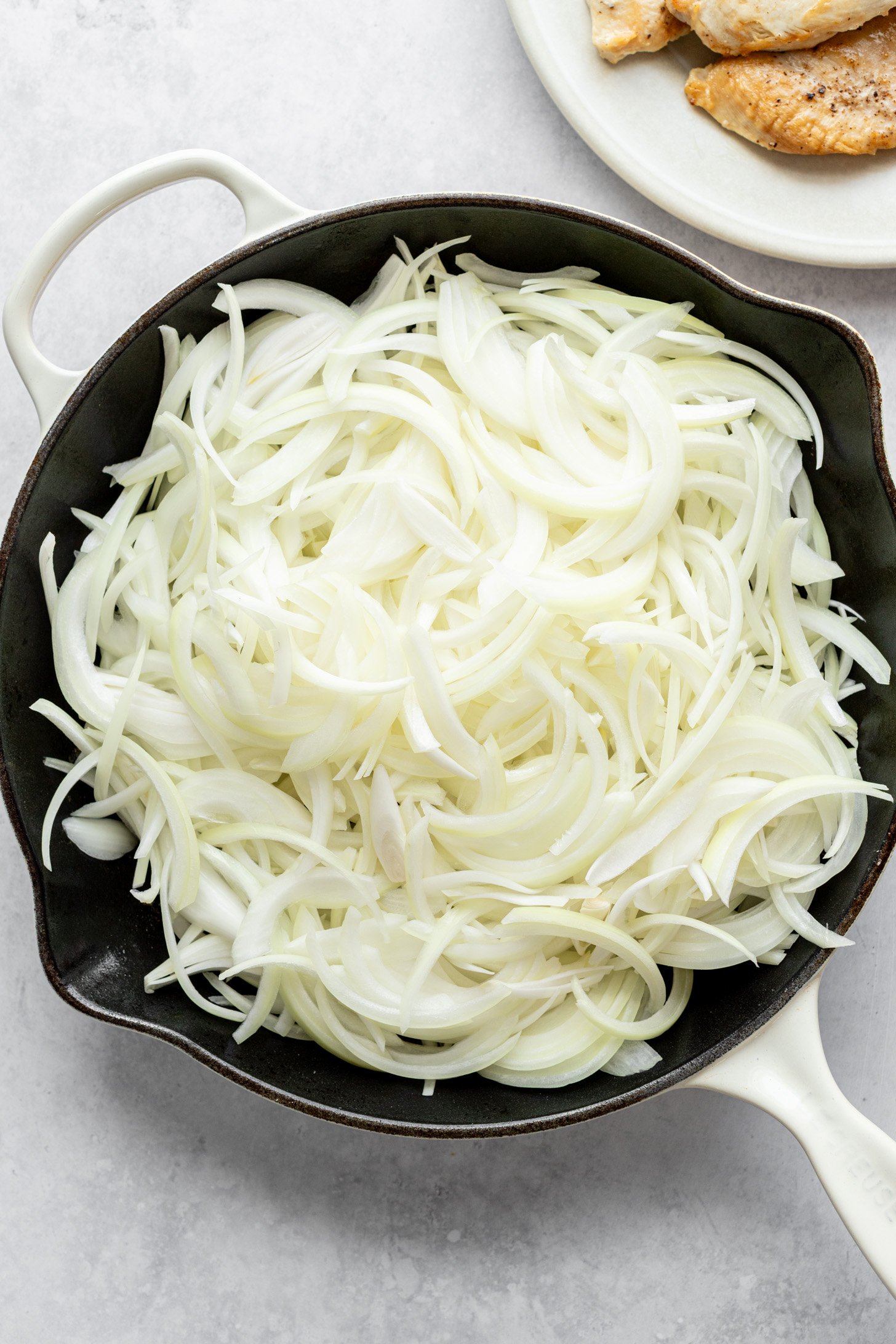 A cast iron skillet filled with raw, sliced white onions. The skillet is sitting on a countertop surrounded by a plate filled with cooked chicken.