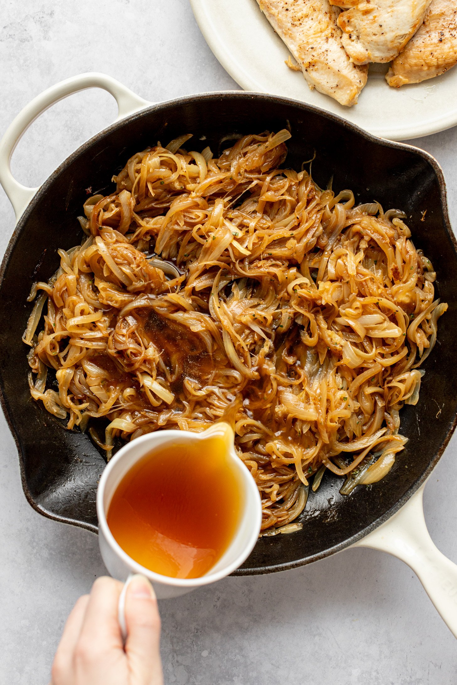 Broth and Worcestershire sauce are being poured into a cast iron skillet filled with caramelized onions. The skillet is sitting on a countertop surrounded by a plate filled with cooked chicken breasts.