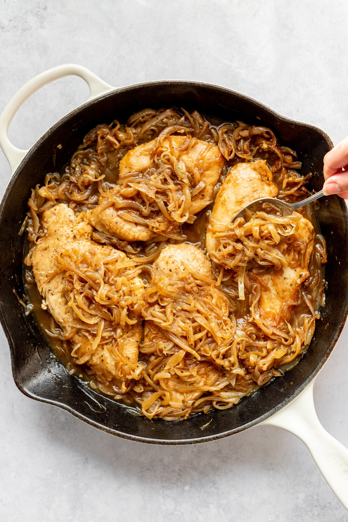 A cast iron skillet filled with caramelized onions and cooked chicken breast being stirred with a spoon. The skillet is sitting on a countertop.