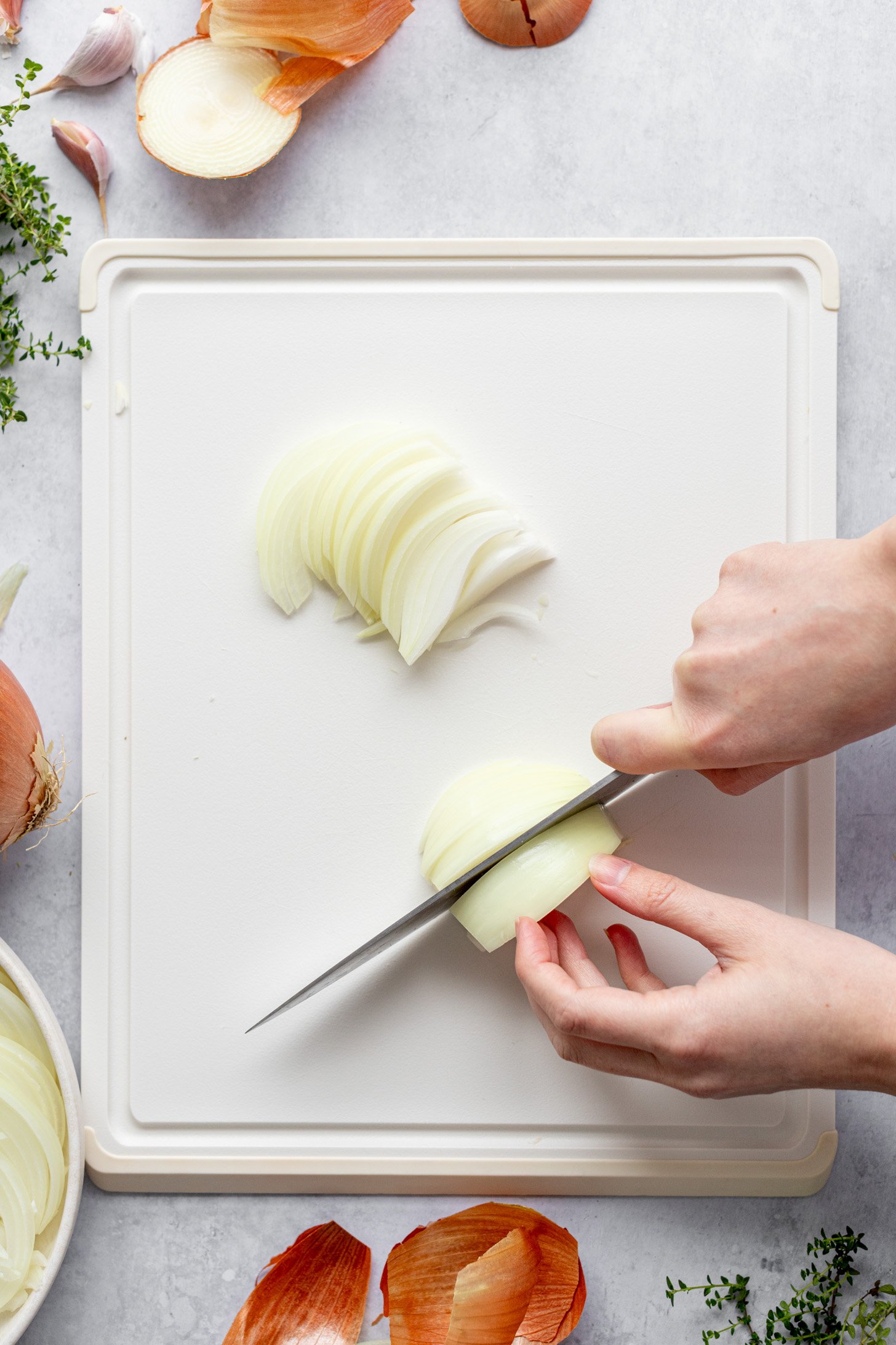 A pair of hands slicing an onion with a knife on a cutting board. The cutting board is on a countertop surrounded by onion peels, discarded pieces of onion, some garlic cloves and some fresh herbs.